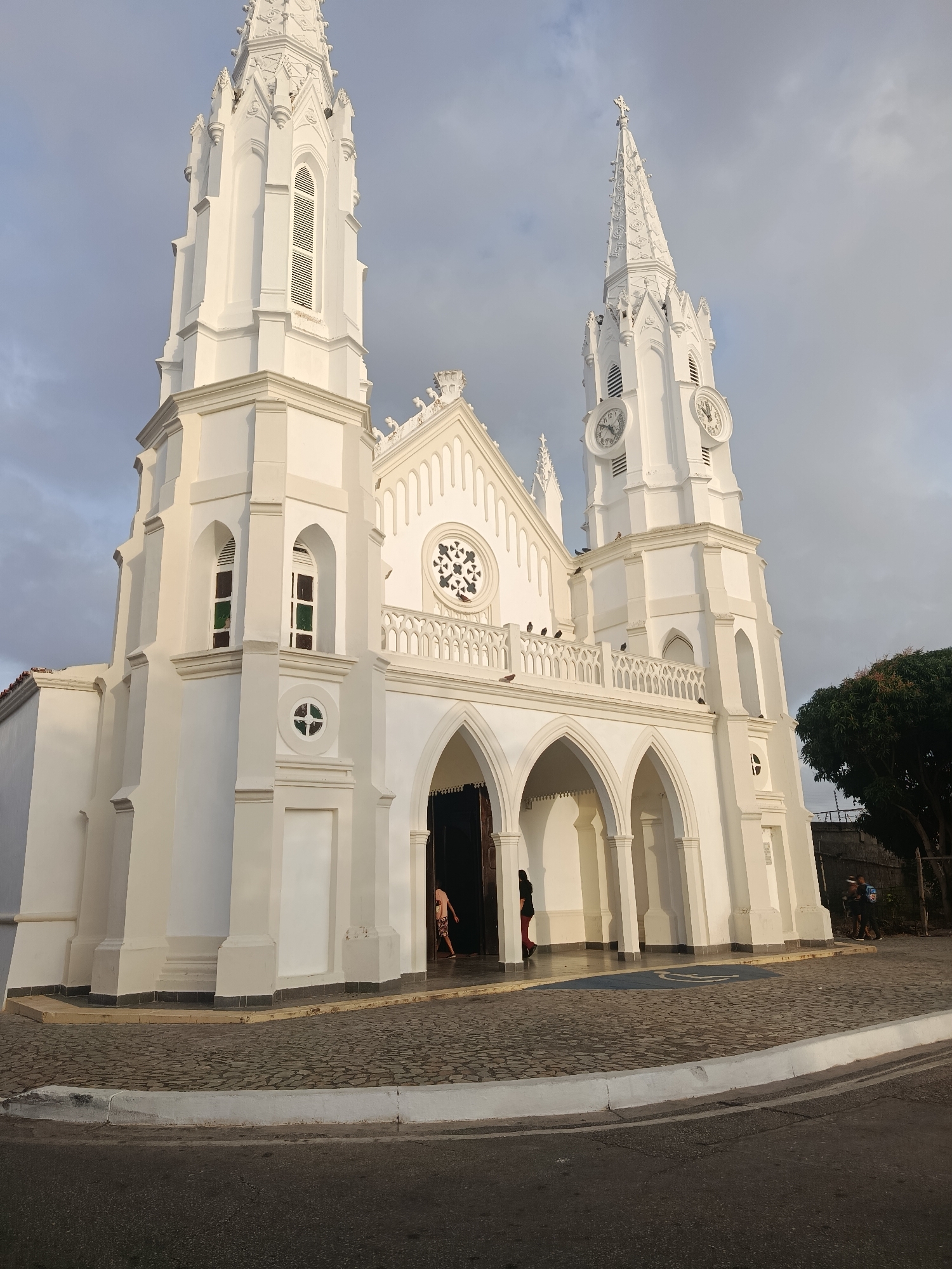 The image shows the neo-Gothic Church of San Juan Evangelista in Juan Griego, Venezuela. The church is predominantly white, featuring two tall towers. The tower on the right contains two clock faces, while the other tower has open spaces. The building has arched entrances and windows. Several people are seen near the entrance to the church. The foreground includes a cobblestone pavement that ends at the roadside. A tree is located to the right of the church. The sky is overcast.