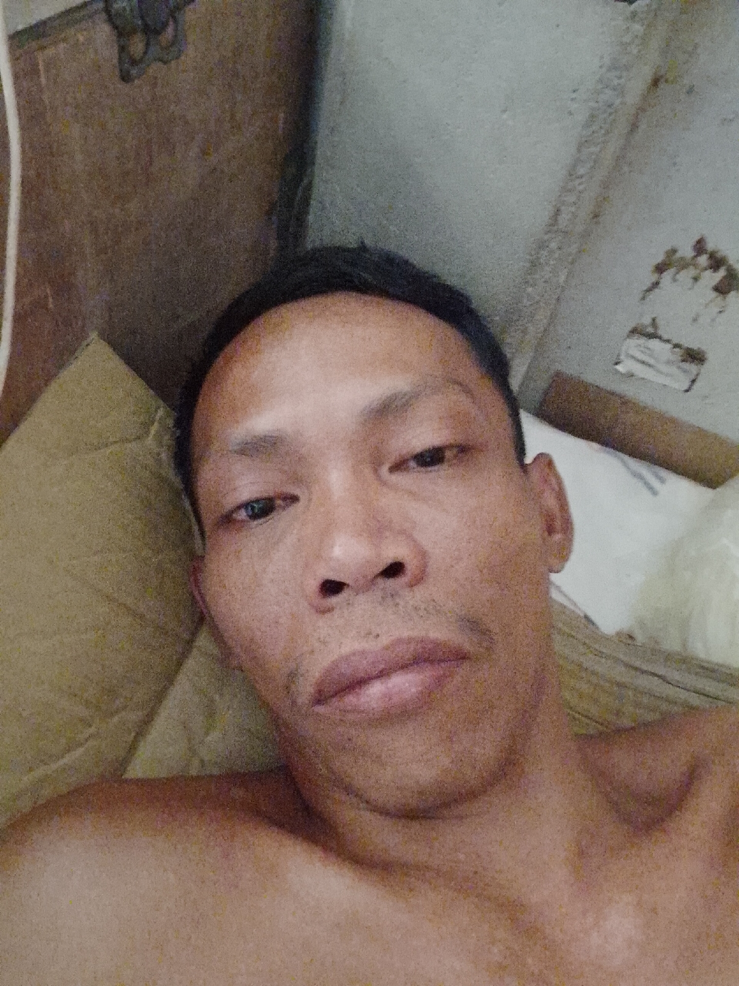 The image shows a selfie of a middle-aged Asian man with short black hair, lying on a tan pillow. The man is shirtless. The background appears to be a wall, part of which is painted white and part is a brown-colored surface. The image quality is low. There is no verifiable information to confirm the location is Bukit Ketok, Indonesia.