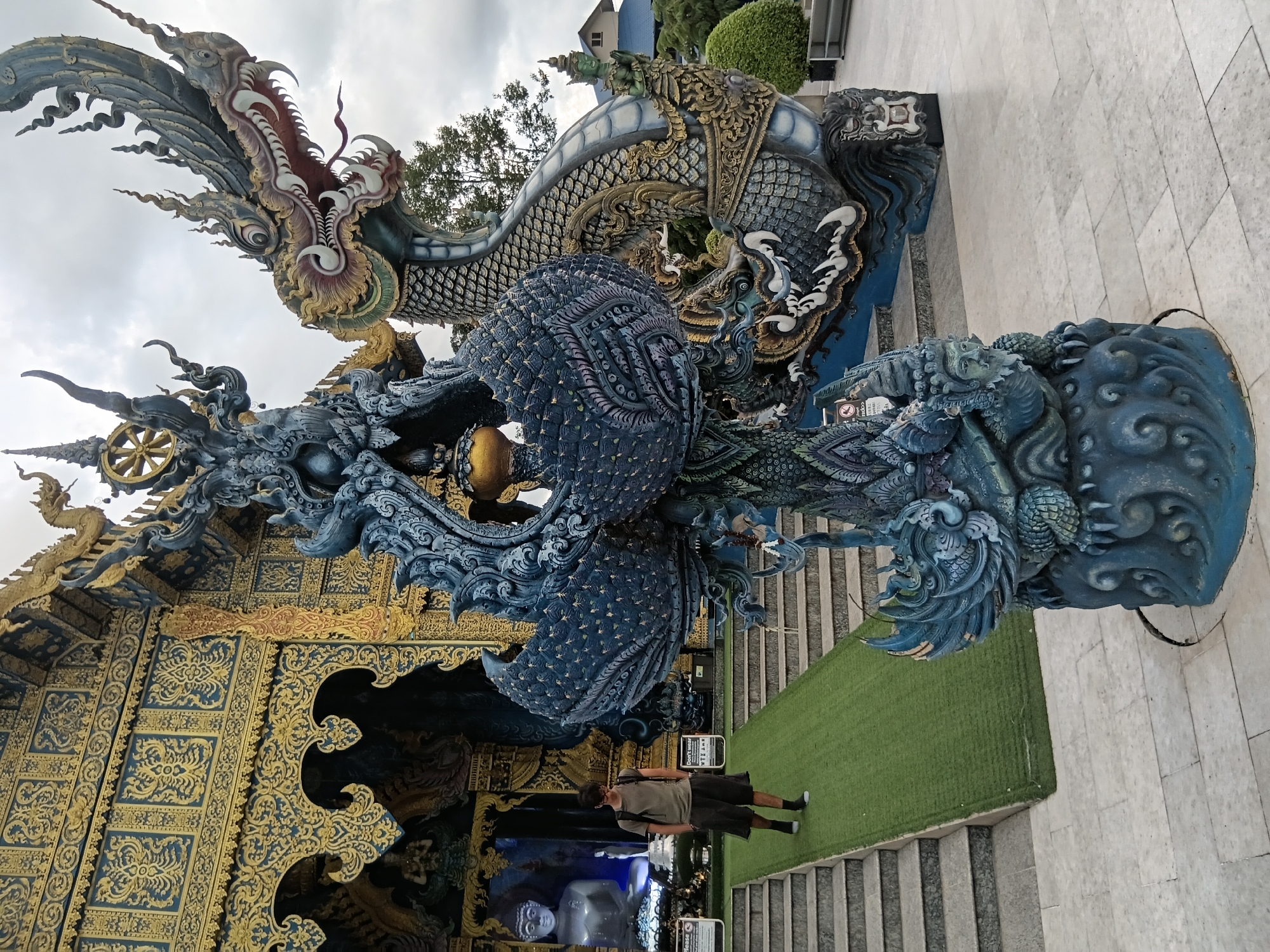 An outdoor, eye-level, medium shot shows the Wat Rong Suea Ten (Blue Temple) in Chiang Rai, Thailand. The temple's ornate, blue and gold facade is visible in the background. In the foreground, a large, blue and gray dragon sculpture with intricate details dominates the scene. To the left of the sculpture, a young man wearing a t-shirt and shorts walks down a green, carpeted ramp adjacent to a set of stone stairs. A large, blue Buddha statue is visible through the temple entrance behind the man. The ground is paved with light-colored tiles. The sky is overcast.