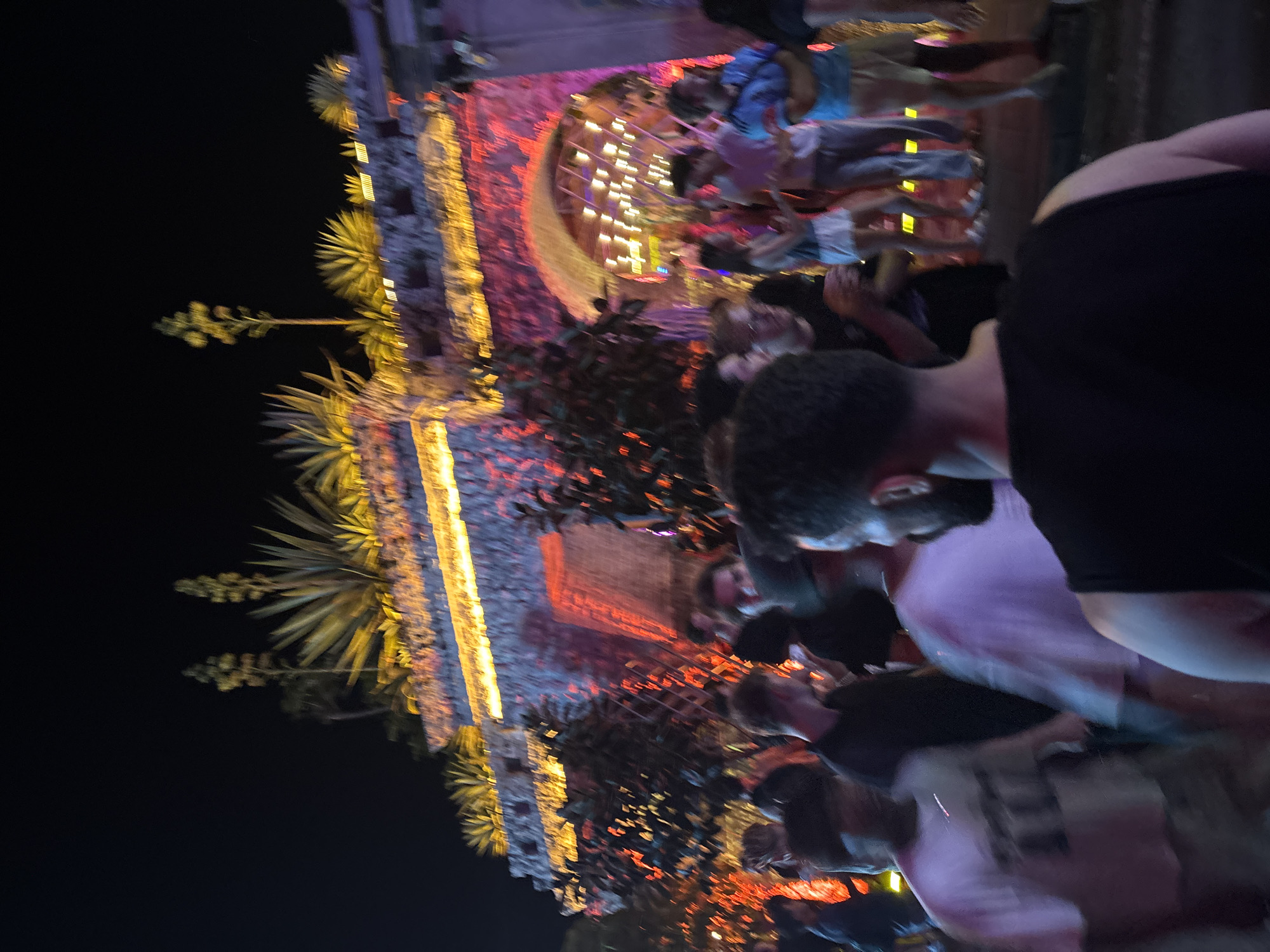 A nighttime shot depicts a crowd of people gathered in front of a stone structure illuminated with vibrant red and yellow lights. The structure features an arched entryway, and its upper sections are adorned with spiky, yellow-hued plants. The scene appears to be an outdoor event, possibly a festival or party, with many individuals blurred due to motion or the photograph's focus. The atmosphere is energetic, suggested by the bright lighting and the presence of a large group. The location is identified as North Kuta, Indonesia.