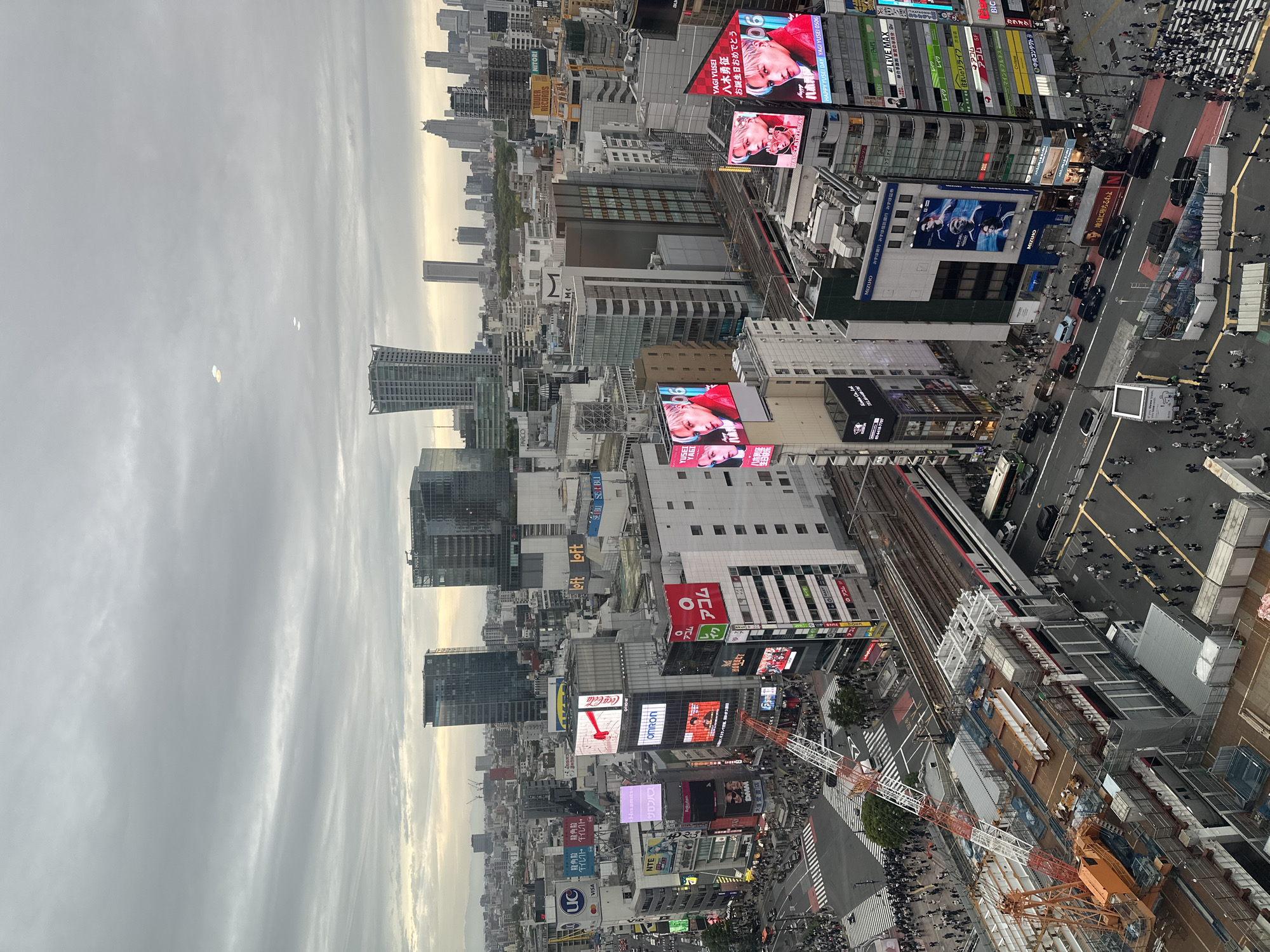 The image presents an aerial view of a bustling urban scene in Shibuya, Japan, under an overcast sky. The environment features high-rise buildings densely packed together, interspersed with large digital billboards displaying advertisements and promotional content. Pedestrians crowd the streets and crosswalks below, indicating high levels of activity. Railway lines and trains are visible among the buildings, suggesting a complex urban infrastructure. Construction is underway, with cranes and construction materials visible, indicating ongoing development or renovation. The overall scene conveys a sense of concentrated commercial and pedestrian activity typical of a major urban center.