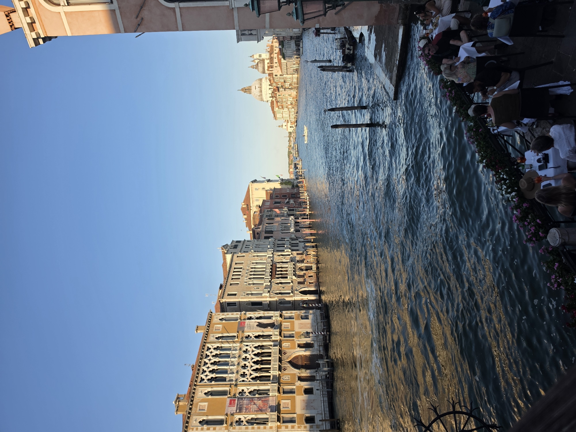 The image depicts a view of the Grand Canal in Venice, Italy, on a clear day with a bright blue sky. In the foreground, a restaurant patio is set up along the canal's edge, with several tables occupied by diners. Some individuals are seen eating, drinking, and conversing. Lush flower boxes with pink and white blossoms line the patio.
The Grand Canal itself is wide and a deep blue, reflecting the sky and the surrounding buildings. The water is slightly rippled, with a boat visible in the distance. On the left side of the canal, a row of historic Venetian buildings with intricate architecture lines the waterfront. These buildings feature ornate windows, balconies, and arched doorways, with banners hanging from some of them.
On the right side of the canal, a building with a pink facade and arched windows is visible, featuring classic Venetian architectural elements. Further down the canal, in the background, the iconic domes of the Basilica di Santa Maria della Salute are prominent, along with other buildings and a boat sailing on the water. The overall scene conveys a vibrant and lively atmosphere characteristic of Venice.