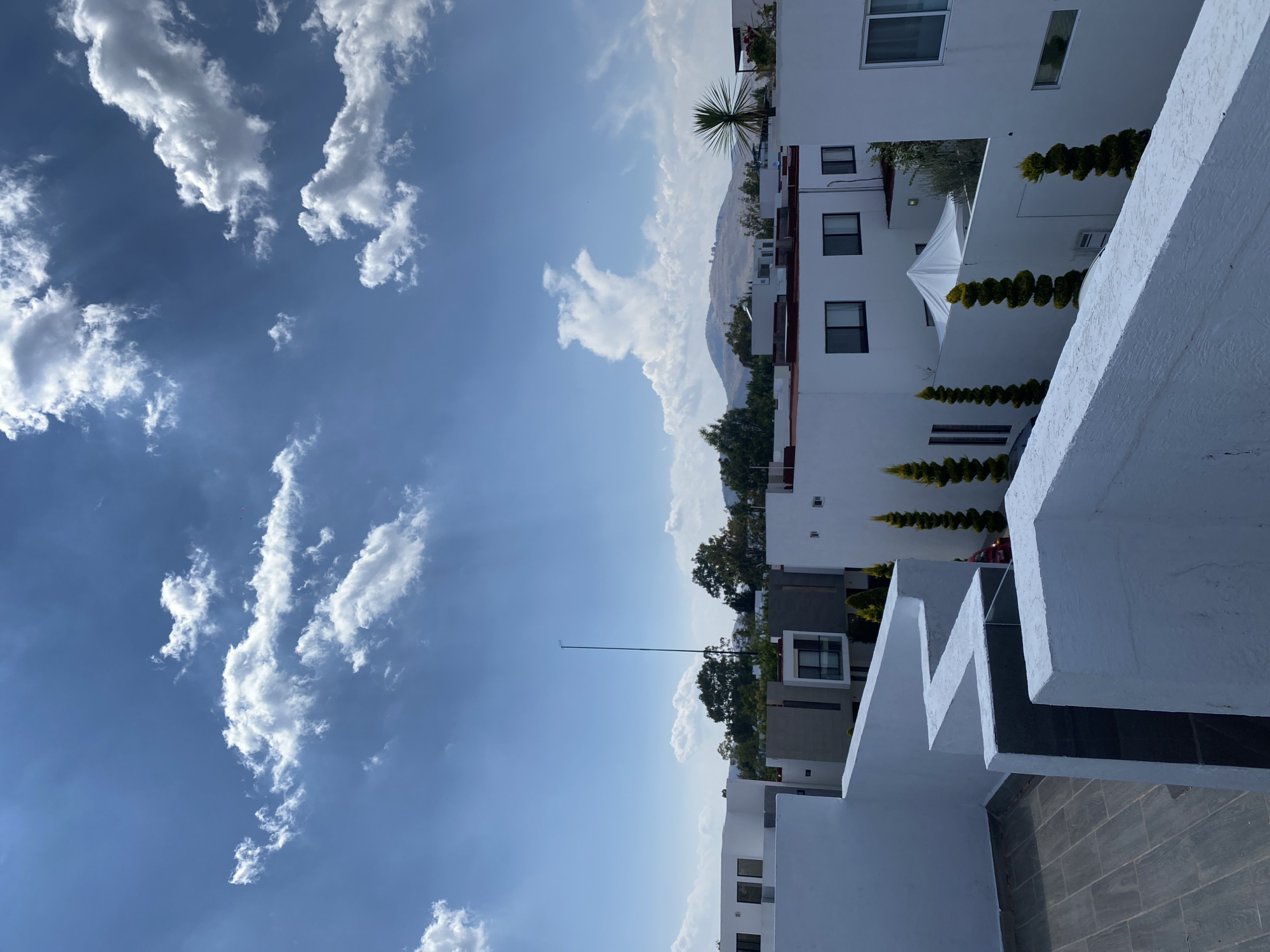 The image shows a view in San Andrés Cholula, Mexico, featuring a bright blue sky with scattered white clouds. White buildings with flat roofs and dark-framed windows are positioned on the right side of the image, with green trees and small trimmed bushes along the building edges. In the distance, a low, brown mountain range is visible. The image is rotated counter-clockwise.