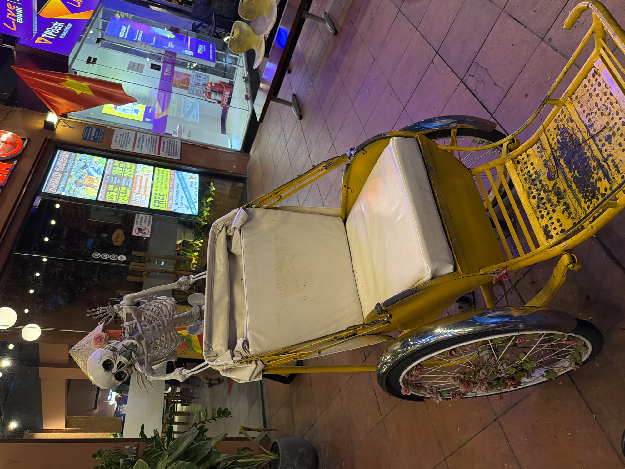 A skeleton wearing a conical hat and adorned with a rose sits in a yellow cyclo, a three-wheeled passenger vehicle common in Vietnam. The skeleton is posed with one hand raised, as if waving. The cyclo is parked on a tiled sidewalk outside a building with large glass windows, reflecting the lights and signage of the shops within. One sign displays prices in Vietnamese Dong, indicating a business related to food or services. A Vietnamese flag hangs from the building. The scene appears to be a street in Nha Trang, Vietnam, possibly decorated for a holiday or event, given the skeleton. The overall atmosphere is a blend of festive decoration and urban commercial activity.