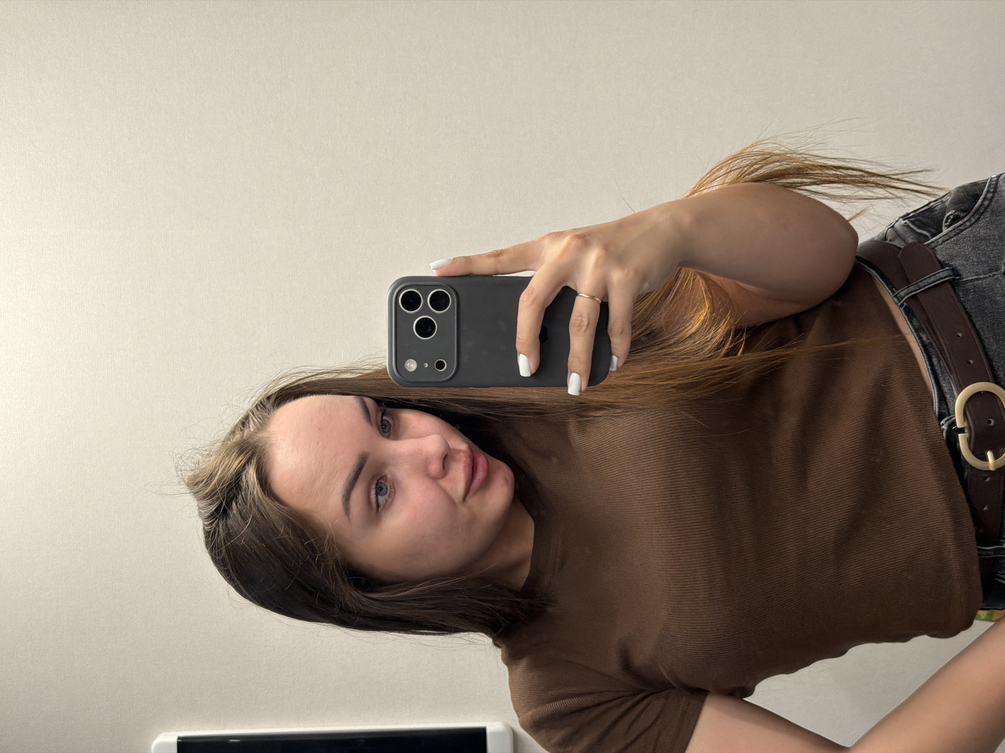 In an indoor setting, a young woman with long, brown hair is taking a selfie in a mirror. She is wearing a brown, ribbed, short-sleeved shirt and dark jeans with a brown belt. Her left hand is resting on her hip, and her right hand is holding a black smartphone up to take the picture. The background is a plain, light-colored wall. There are no discernible landmarks or contextual clues to suggest the location is in Kamyennoye, Russia.