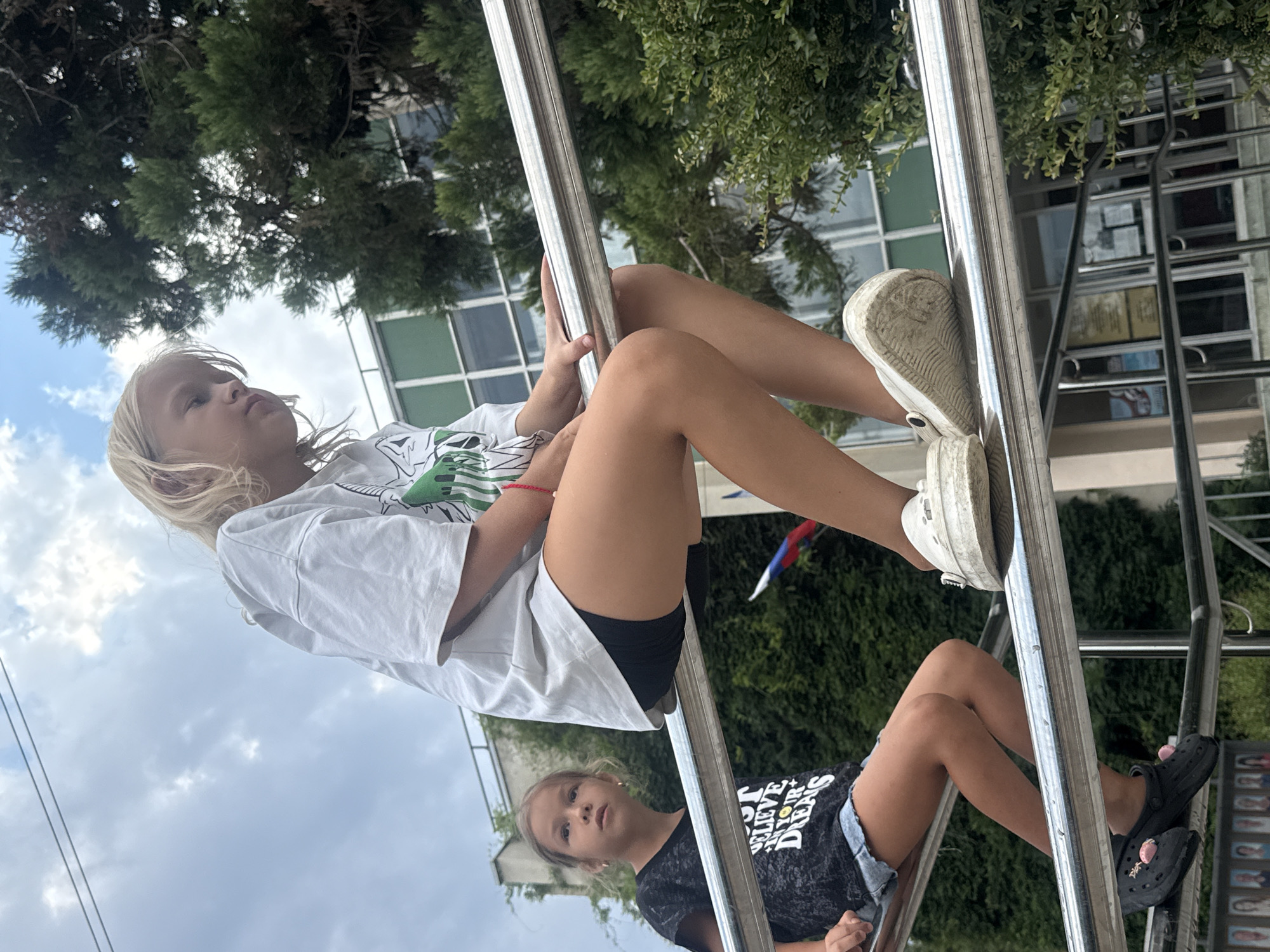 Two young girls, one with blonde hair and the other with brown hair, are sitting on a metal railing. The blonde girl, in the foreground, is wearing a white t-shirt and black shorts. The girl with brown hair, in the background, is wearing a black t-shirt and denim shorts. They appear to be in an outdoor urban setting with trees and a building visible behind them. The blonde girl is looking to her right, while the brown-haired girl is looking forward. A small flag with blue, white, and red stripes is visible behind the brown-haired girl. The overall scene suggests a casual outdoor activity.