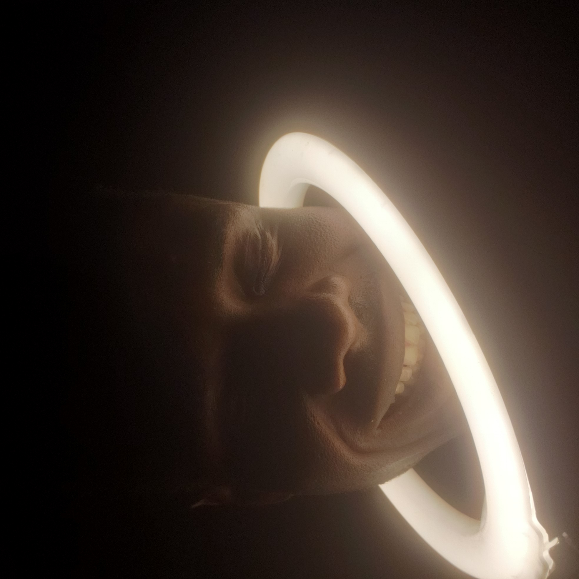 A close-up, low-key image features a person's face partially illuminated by a ring light. The person is smiling, and their teeth are visible. The background is dark, providing a high contrast with the illuminated face and ring light. The location, Narayi, Nigeria, is not evident in the visual content of the image. There are no discernible objects or actions beyond the person's smile and the presence of the ring light.