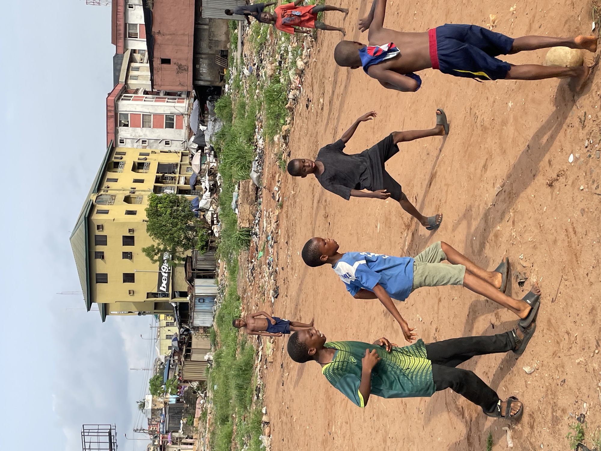 A group of young boys are playing football in a dirt field in Onitsha, Nigeria. The background consists of buildings and a large accumulation of garbage. The boys are wearing various casual clothes, including shorts, t-shirts, and sandals. They are in motion, appearing to kick or run after a football. One boy is shirtless. The sky is overcast, and the urban landscape shows signs of poverty and environmental neglect.