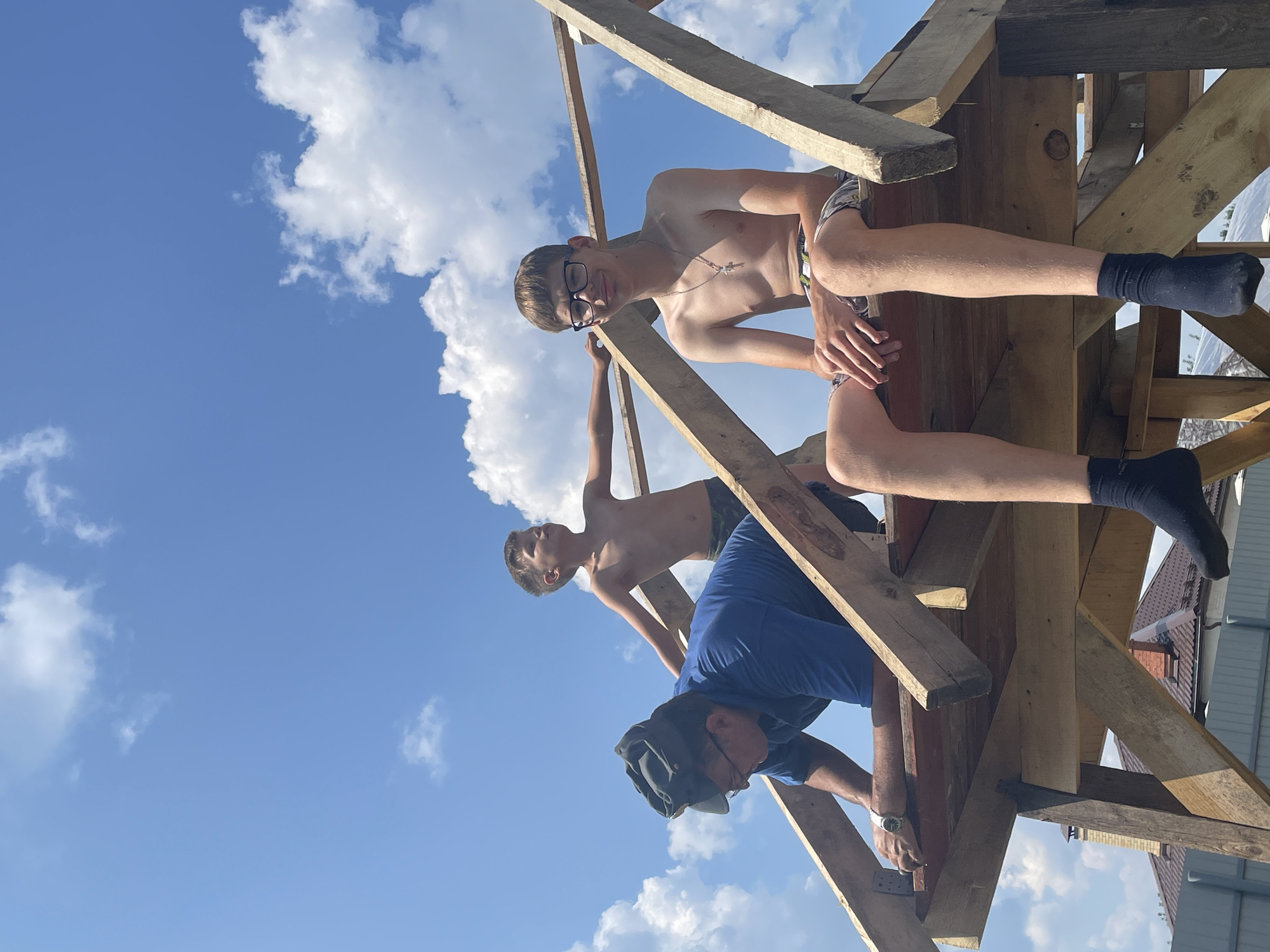 Three individuals are positioned on a wooden structure, likely under construction, against a bright blue sky with scattered white clouds. The person in the foreground, on the right, is a young male wearing glasses and swim trunks, sitting with his legs extended. He is bare-chested and wears a silver necklace with a cross pendant and dark socks. To his left and slightly behind him, another young male, also bare-chested and wearing swim trunks, is leaning forward, with his arms outstretched. In the center-left of the image, an adult male wearing a blue shirt and a cap is bent over, appearing to be working on the structure. His hands are visible, and he wears a watch on his left wrist. The wooden structure consists of beams and planks, suggesting it is a roof or part of a building frame. In the background, rooftops of houses and fencing are visible, indicating an outdoor, possibly residential, setting. The overall lighting suggests a sunny day. The specific location in the city of Маминское, Russia is not discernible from the visual information alone.