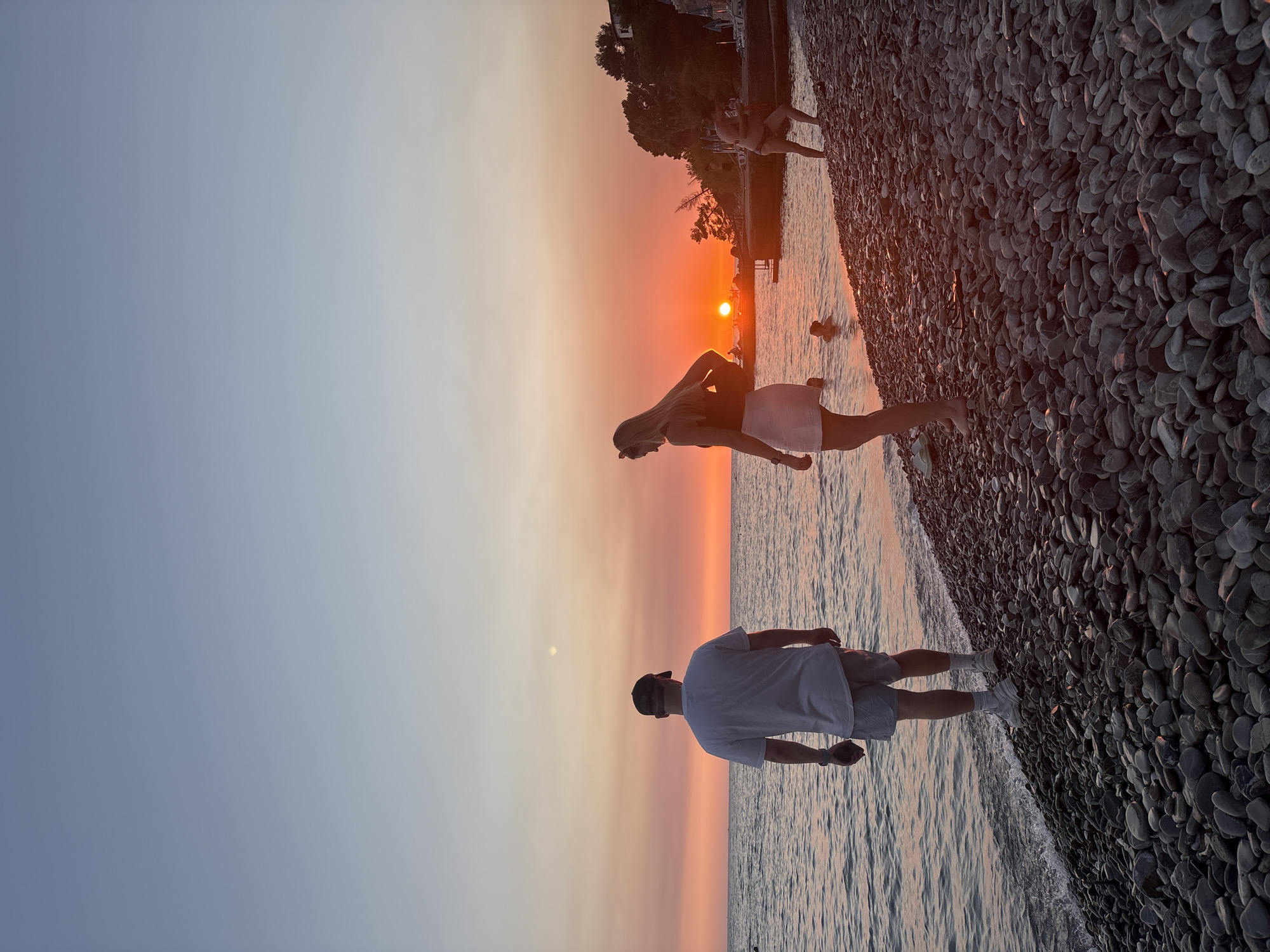 A wide shot of a pebble beach in Bar, Montenegro, at sunset. The sky is a gradient of pale blue to orange, with the sun low on the horizon, casting a warm glow over the scene. The sea is calm and reflects the sky.
In the foreground, a man wearing a white t-shirt and shorts stands facing the sea, his back to the camera. To his right, a woman in a white skirt and a darker top stands also facing the sea, with her back to the camera. Further to the right, in the water, a person is partially submerged, and another person stands on the rocky shore.
The beach is covered in dark grey pebbles. Trees and buildings are visible on a hill in the background. The overall impression is of a peaceful evening scene by the sea.