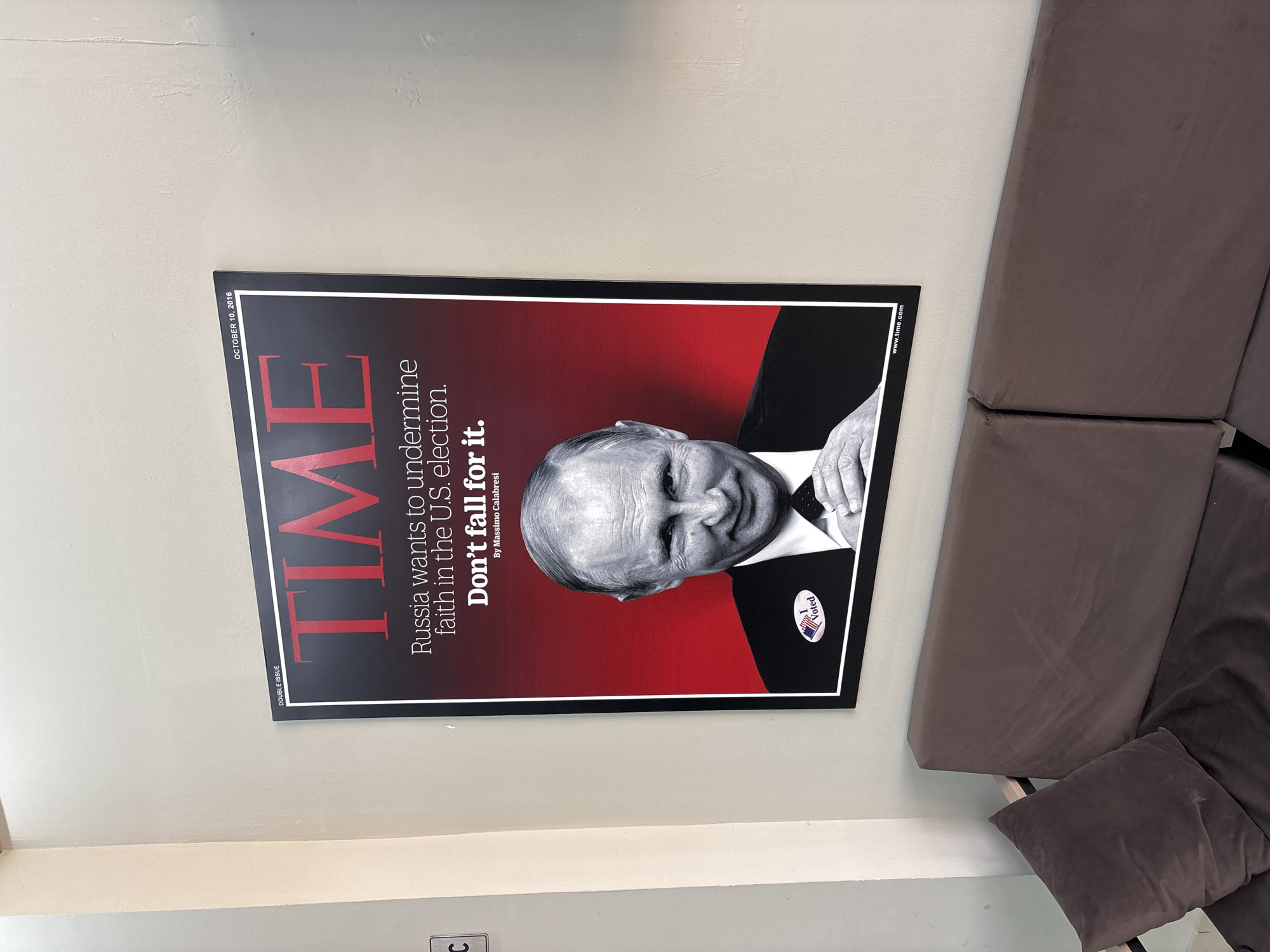 The image shows a framed magazine cover hanging on a wall next to a sofa. The magazine is TIME, dated October 10, 2016. The text on the cover reads, "Russia wants to undermine faith in the U.S. election. Don't fall for it." There is a black and white photograph of Vladimir Putin on the cover, with a "I Voted" sticker on his lapel. The sofa is brown and has a throw pillow. The location or the city of Chaikovsky, Russia, is not explicitly confirmed from the content of this image.