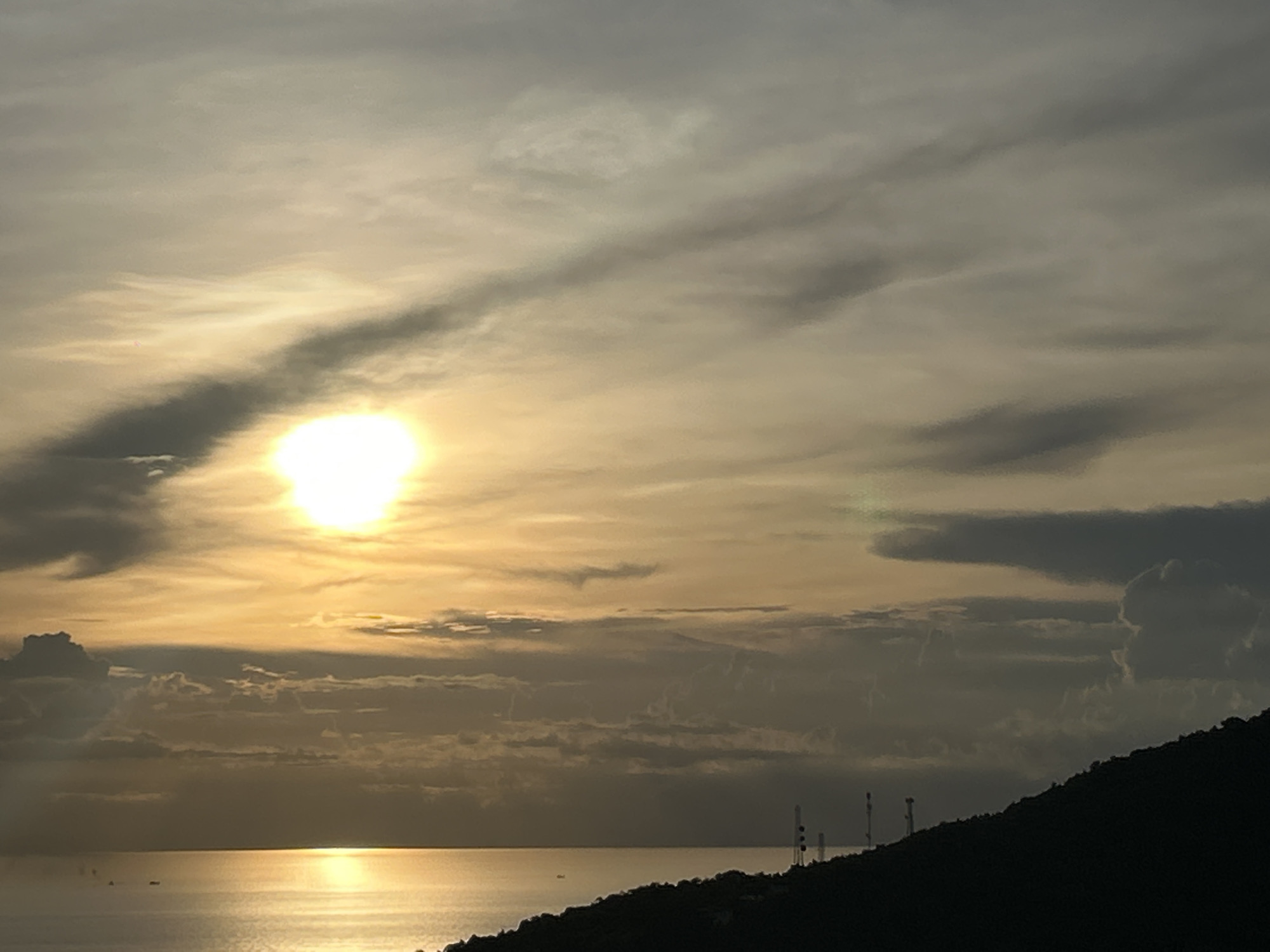 The image depicts a sunset over the ocean, likely in Koh Phangan, Thailand. The sun, a bright orb, is partially obscured by clouds, casting a golden glow on the water. The sky is a mix of warm yellow and grey hues, with dramatic cloud formations stretching across the horizon. In the foreground, a dark silhouette of a hillside slopes down to the sea, dotted with what appear to be telecommunication towers. A few small boats are visible on the calm water. The overall mood is serene and peaceful, evoking a sense of a beautiful tropical evening. The time of day is clearly sunset, given the sun's position and the warm, diffused light. There is no visible text in the image.