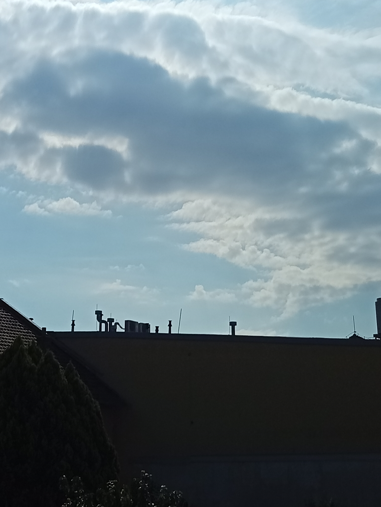 The image depicts a view of rooftops against a cloudy sky. The foreground is dominated by the dark silhouettes of trees and buildings. On the rooftops, various ventilation pipes and possibly air conditioning units are visible. The sky is filled with layered clouds, with patches of lighter blue sky showing through. The scene is set in the city of Tišnov, Czechia.