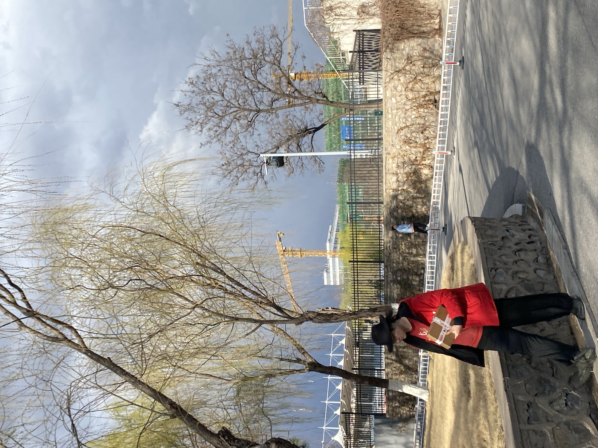 In Zhongtun, China, a person wearing a red jacket and black pants stands on a stone ledge. The individual holds a cardboard box wrapped with white tape. A black hat rests atop their head, and a black jacket is worn underneath the red one. Behind them is a street lined with a stone wall and metal fences, with trees present on both sides. A construction crane is visible in the background against a cloudy sky. A ladder is extended alongside the road.