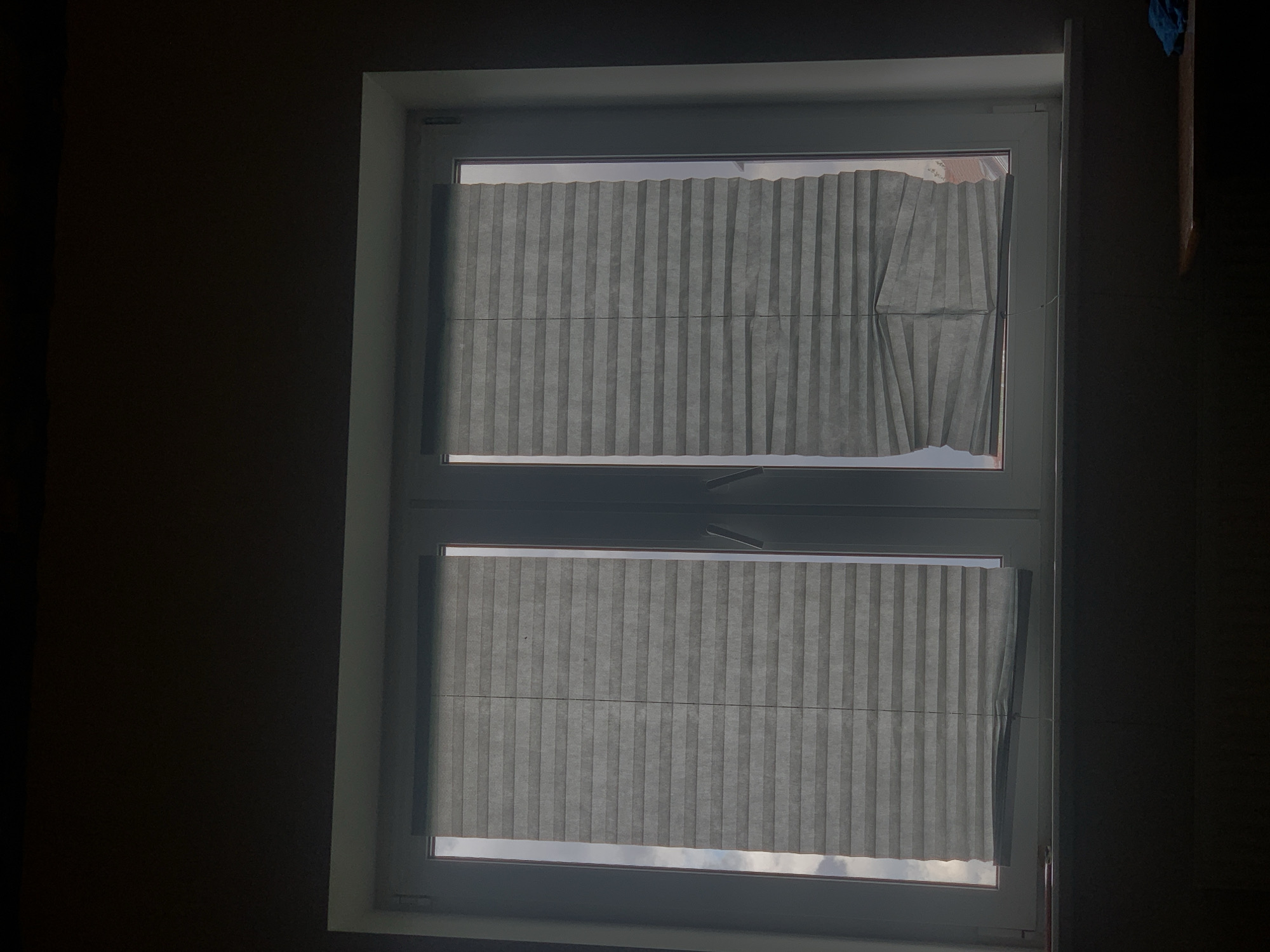 A close-up, dimly lit shot shows a window with two pleated blinds, partially covering the panes. The blinds are gray and appear to be made of a fabric-like material. The left blind is fully extended, while the right blind is partially lowered and appears creased or uneven. The window frame is white. Outside the window, the sky is visible, with some clouds and hints of buildings. The foreground is dark, suggesting the photo is taken from inside a room looking out. The image's overall tone is muted and slightly somber. No people are visible.