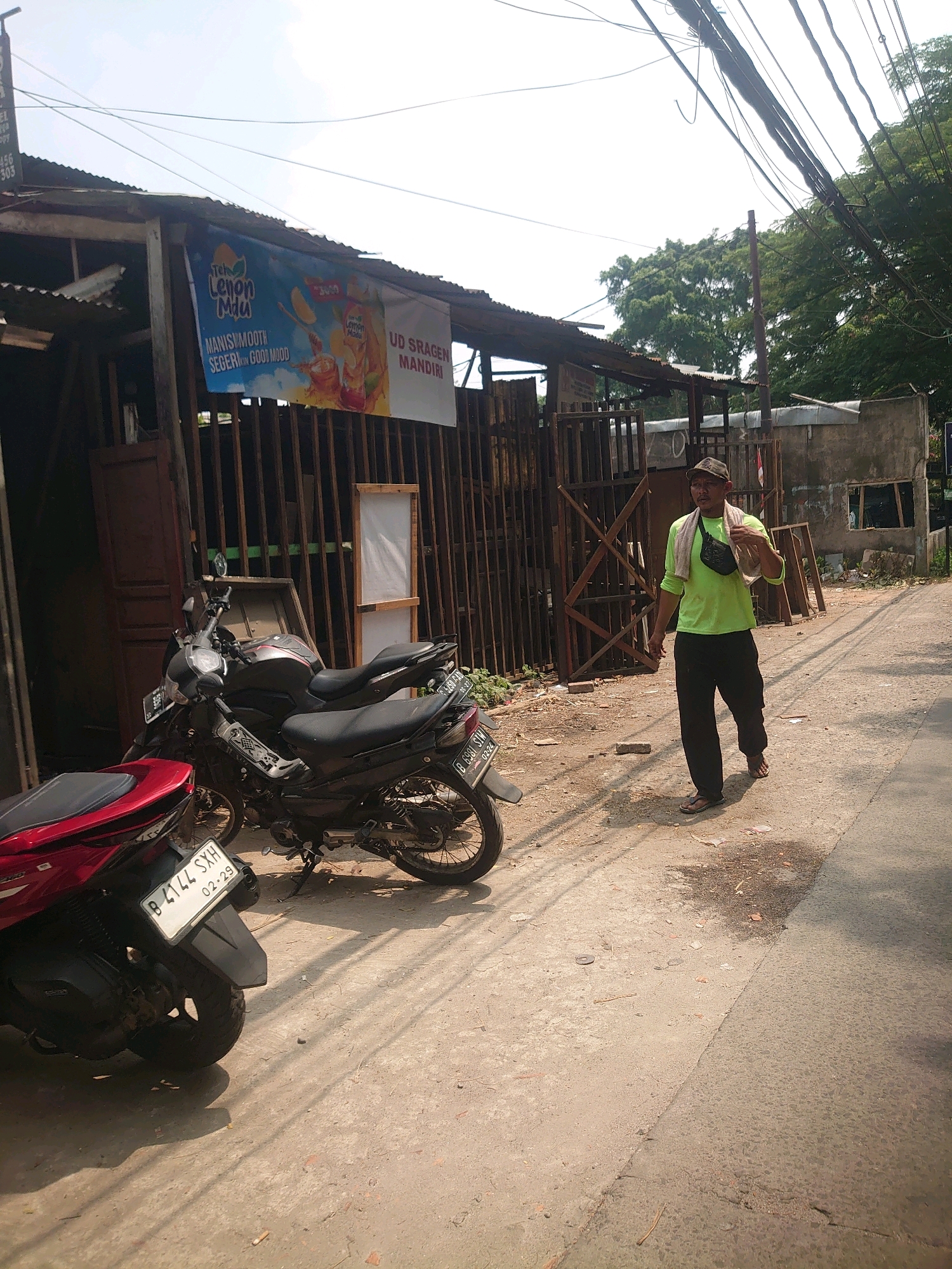 The image captures a sun-drenched street scene in Depok, Indonesia, with a man walking along the right side of the frame. He is wearing a lime green long-sleeved shirt, black pants, brown flip-flops, a dark fanny pack, and a towel draped over his left shoulder. A baseball cap is on his head. In the background, a modest, open-fronted building with a corrugated metal roof stands, displaying a large banner advertising "Teh Lemon Muu" and "UD SRAGEN MANDIRI". Several motorcycles are parked in front of the building, including a red scooter in the foreground and two black motorcycles behind it. The ground is a mix of dirt and asphalt, with some debris scattered around. Overhead, a complex network of electrical wires crisscrosses the sky. The overall environment suggests a commercial or residential area in a developing part of the city.