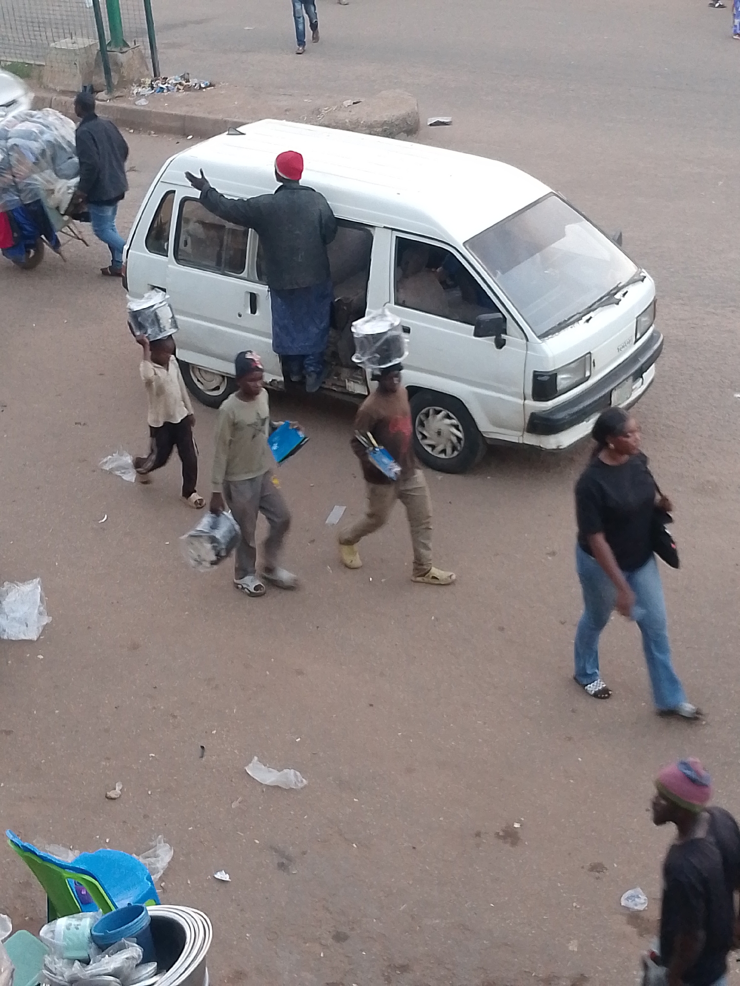 In Bukuru, Nigeria, on an unpaved street, a white minivan is parked. A person wearing a red cap and a blue skirt is standing in the open side door of the minivan, with their arm raised. Several children are walking away from the minivan, each carrying a bucket on their head or in their hands. One child is carrying a blue folder. A woman in a black shirt and blue jeans is walking away from the minivan. Another person, wearing a pink and blue hat, is walking in the foreground. The ground is littered with trash and plastic bags. A wheelbarrow loaded with sacks is to the left of the minivan, and a man in a dark jacket is pushing it.