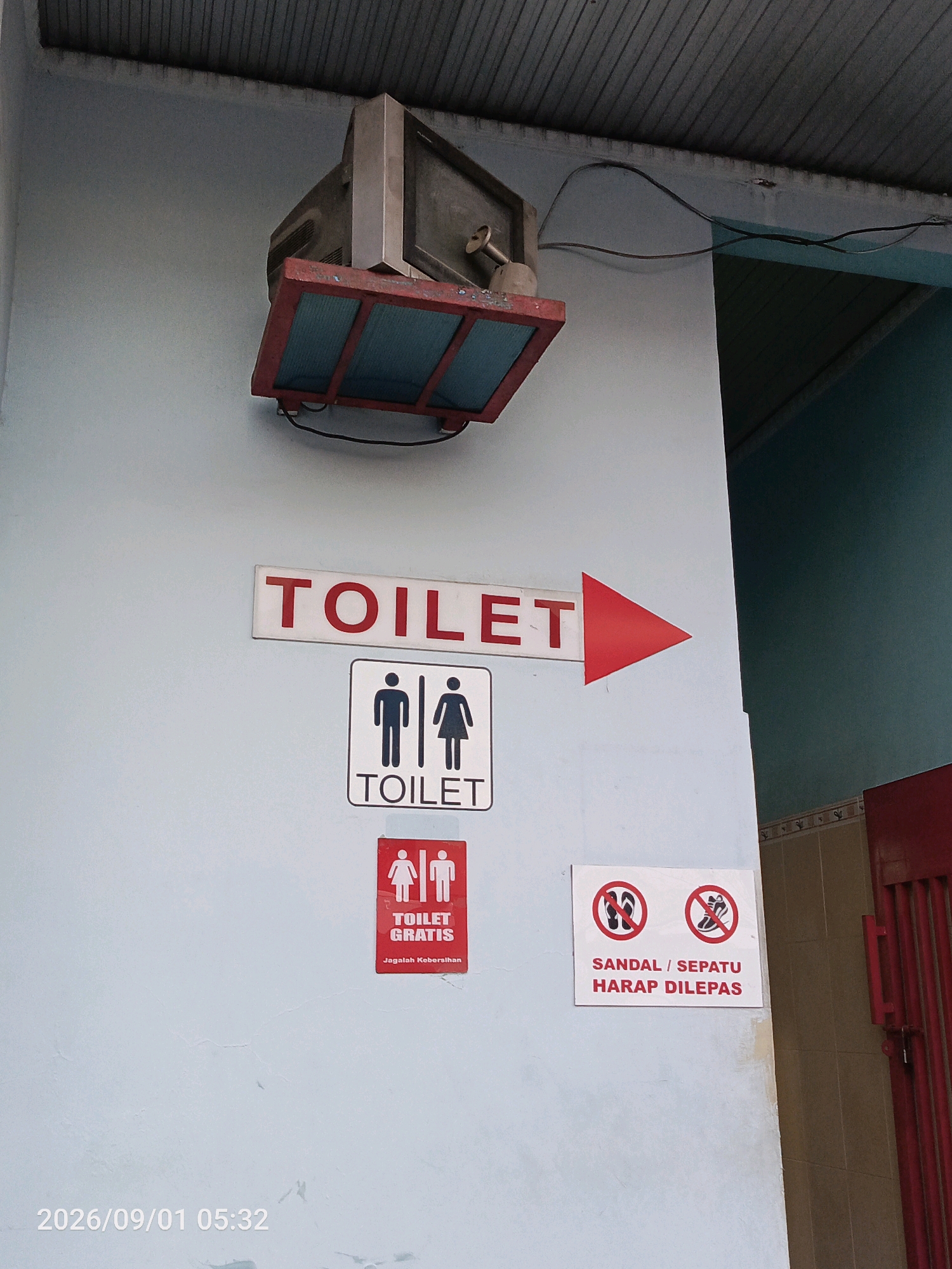 The image shows a light blue wall with several signs. A large red and white sign with the word "TOILET" and a red arrow points to the right. Below this, a black and white sign displays icons of a man and a woman with the word "TOILET" below. To the left and slightly below this, a red sign with white figures shows a man and woman, with "TOILET GRATIS" and "Jagalah Kebersihan" written below. To the right, a white sign with two red circles containing crossed-out sandals and shoes, with the text "SANDAL / SEPATU HARAP DILEPAS" below. A ceiling-mounted object, possibly a speaker or light, is visible at the top. The lower left corner of the image has a timestamp: "2026/09/01 05:32". The image does not contain any explicit information about its location in Gladak Anyar, Indonesia.