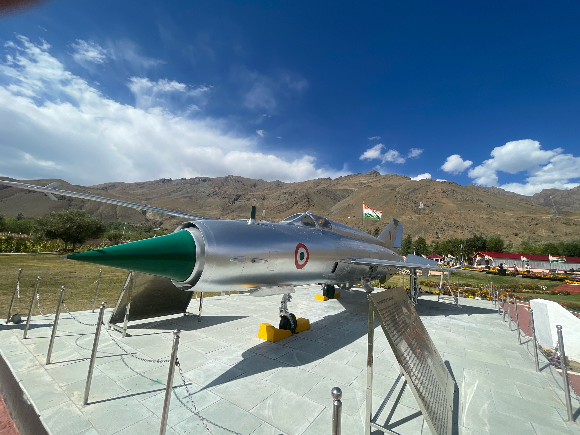 A silver jet fighter, identified by the Indian flag and roundel insignia, is displayed on a paved platform in Bhimbat, India. The aircraft, a Mikoyan-Gurevich MiG-21 according to the informational display, is positioned with its nose cone pointed left and its landing gear down, supported by yellow blocks. The scene is outdoors, with a clear blue sky and scattered white clouds above. In the background, a range of arid, rocky mountains rises, with some sparse vegetation and trees visible at their base. Several Indian flags are displayed on poles throughout the grounds, and a chain-link fence surrounds the aircraft. The paved area features metallic stanchions with chains, guiding visitors around the exhibit. An informational plaque stands in front of the jet, detailing its history. The overall environment suggests a public memorial or museum site.