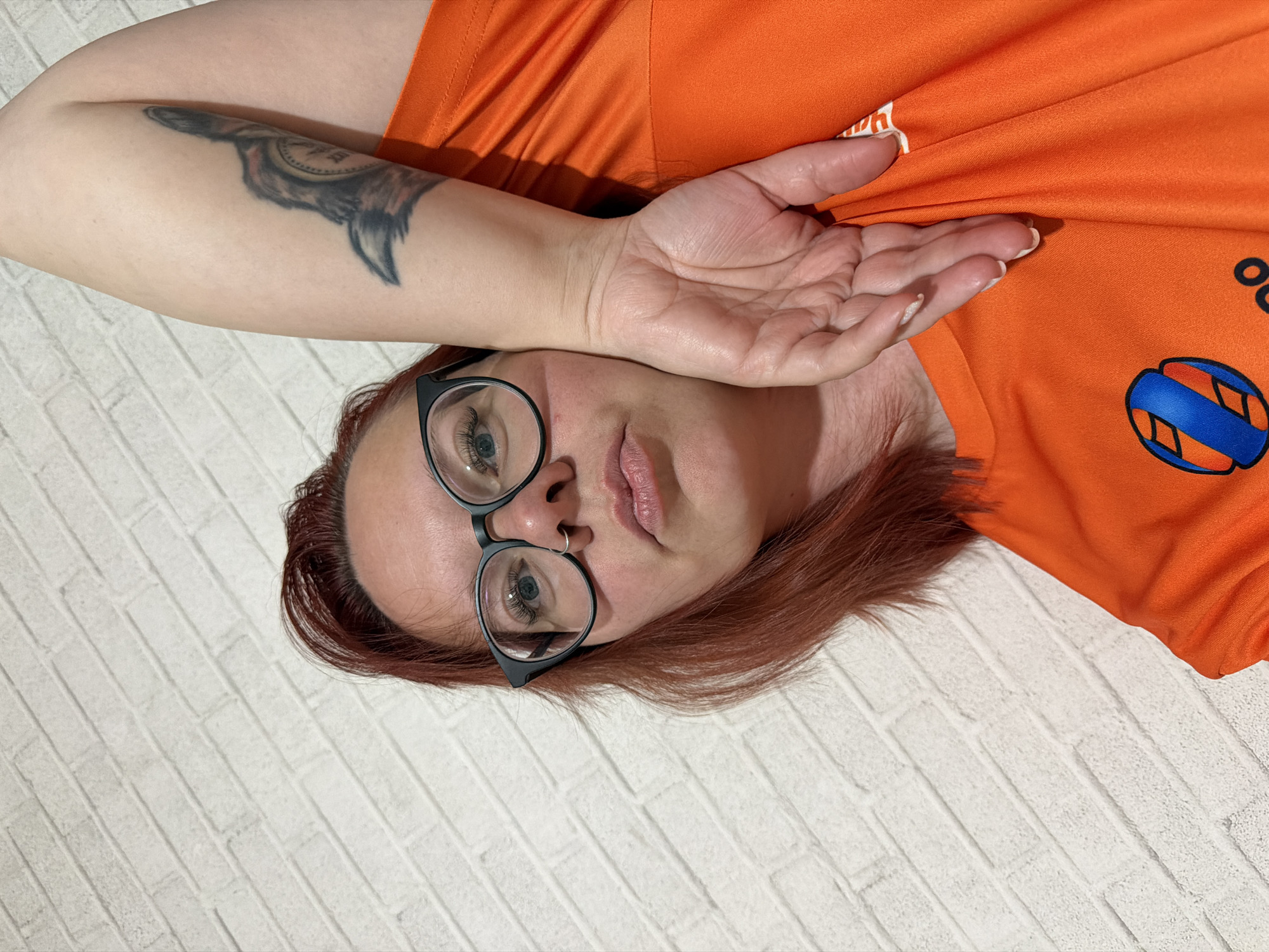 A red-haired woman with glasses and a nose ring is lying on a white brick-patterned surface. She is wearing an orange shirt with a blue and orange design on the right side of the chest. Her left arm is bent with a tattoo visible. Her hand is near her forehead. The location appears to be indoors, but there is no specific information to confirm the location in the city of Chaikovsky, Russia.