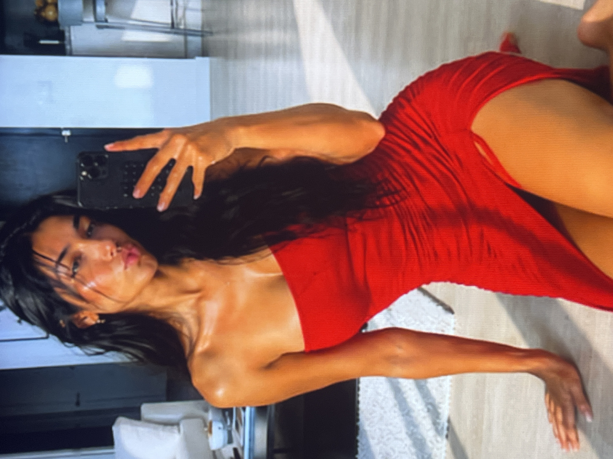 A young woman with long, dark hair is kneeling on a floor, taking a mirror selfie with her phone. She is wearing a strapless red dress with a high slit. The background appears to be an indoor setting, possibly a living room or studio apartment, with furniture and kitchen elements visible. The lighting suggests it is daytime, with sunlight casting shadows on the floor.