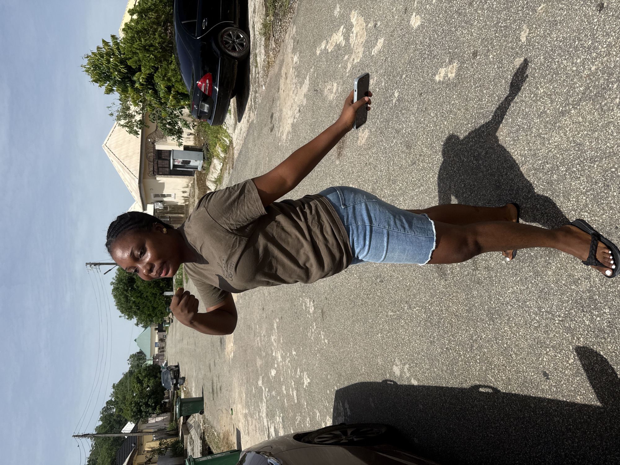 A woman is standing on a paved road outdoors in Abuja, Nigeria. She is holding a smartphone in her outstretched right hand, seemingly taking a photo or video of herself. She is wearing a brown t-shirt and denim shorts, with black sandals on her feet. A black car is parked nearby, partially visible in the background and foreground. The environment includes buildings, trees, and other vehicles along the road, under a bright sky.