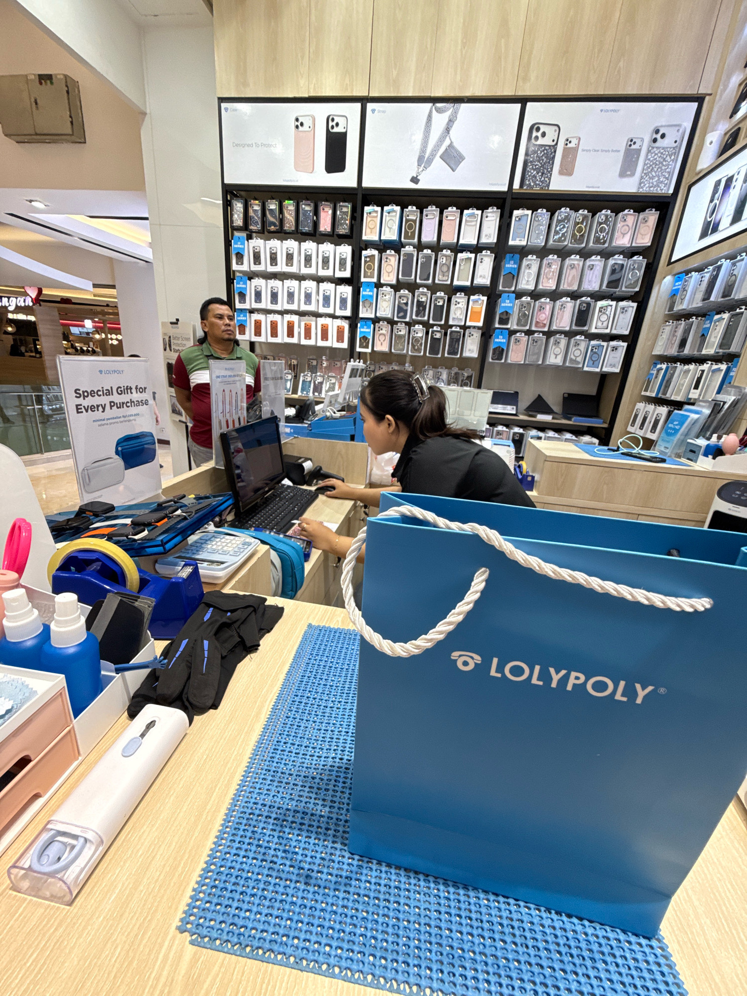 In a brightly lit retail store in Surabaya, Indonesia, a male employee stands behind a counter, looking at a female customer. The store specializes in phone accessories, with a wall display showcasing a variety of phone cases. The counter is cluttered with items, including a laptop, calculator, and various phone accessories. A large blue shopping bag with the logo "LOLYPOLY" is prominently displayed in the foreground. The overall scene suggests a typical retail transaction in a mobile phone accessory shop.