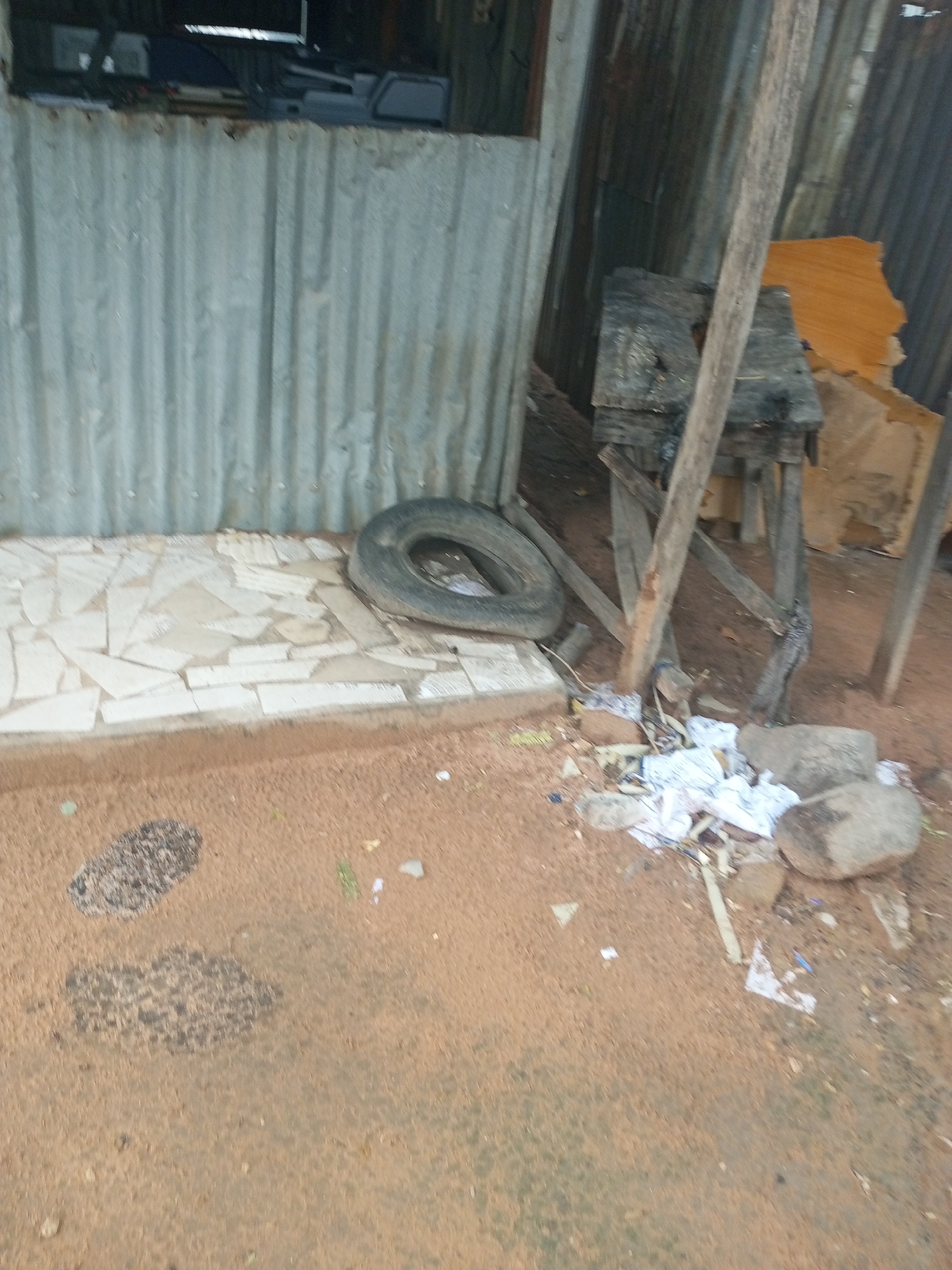 The image depicts an outdoor scene with a corrugated metal wall in the background, possibly part of a makeshift structure or shed. In front of the wall, a rough, tiled surface is visible, upon which a deflated tire lies. The ground in front of the tiled area is sandy and reddish-brown, with scattered debris including white paper, rocks, and possibly some organic matter. A wooden structure, appearing to be a table or workbench supported by angled wooden legs, stands to the right of the tire. This structure is weathered and rustic, with visible grain and imperfections in the wood. A tall, rough wooden post is positioned in front of the wooden structure, leaning slightly. Two dark, textured shapes are visible on the sandy ground to the left, resembling footprints or stains. The overall impression is one of an unkempt outdoor area, potentially a workshop or storage space, in a semi-arid or dusty environment. There is no visible text in the image. The lighting suggests it is daytime. The scattered debris and weathered materials indicate a utilitarian or perhaps abandoned setting.