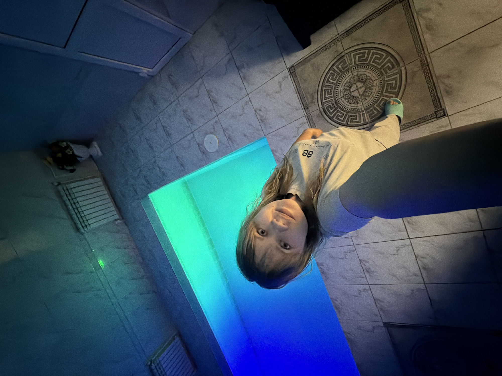 The image depicts a low-angle selfie of a woman taken indoors, likely in a pool or spa area, in the city of Chaikovsky, Russia. A Caucasian woman with shoulder-length brown hair and bangs, wearing a light-colored t-shirt with a visible "BB" logo and light-colored pants, looks directly at the camera with a subtle smile. Her left foot, partially visible, is adorned with a light blue slide sandal.
Behind and to the left of the woman, a rectangular pool or water feature is brightly illuminated by internal LED lighting, displaying a vibrant blue on the left and a luminous green on the right. The floor is covered with light-toned, possibly marble-patterned, square tiles. A prominent circular decorative tile with a Greek key pattern and a central emblem is visible on the right foreground.
The walls and ceiling are light-colored and appear textured. In the upper left, a rectangular ceiling vent is present. On the upper right wall, a white radiator is mounted, with a small, glowing green light positioned above it. A white vase holding artificial yellow and orange flowers is situated in the upper right corner, adjacent to a closed white door with a visible frame. The primary illumination in the scene originates from the colored pool lights, casting distinct blue and green tones across the environment.