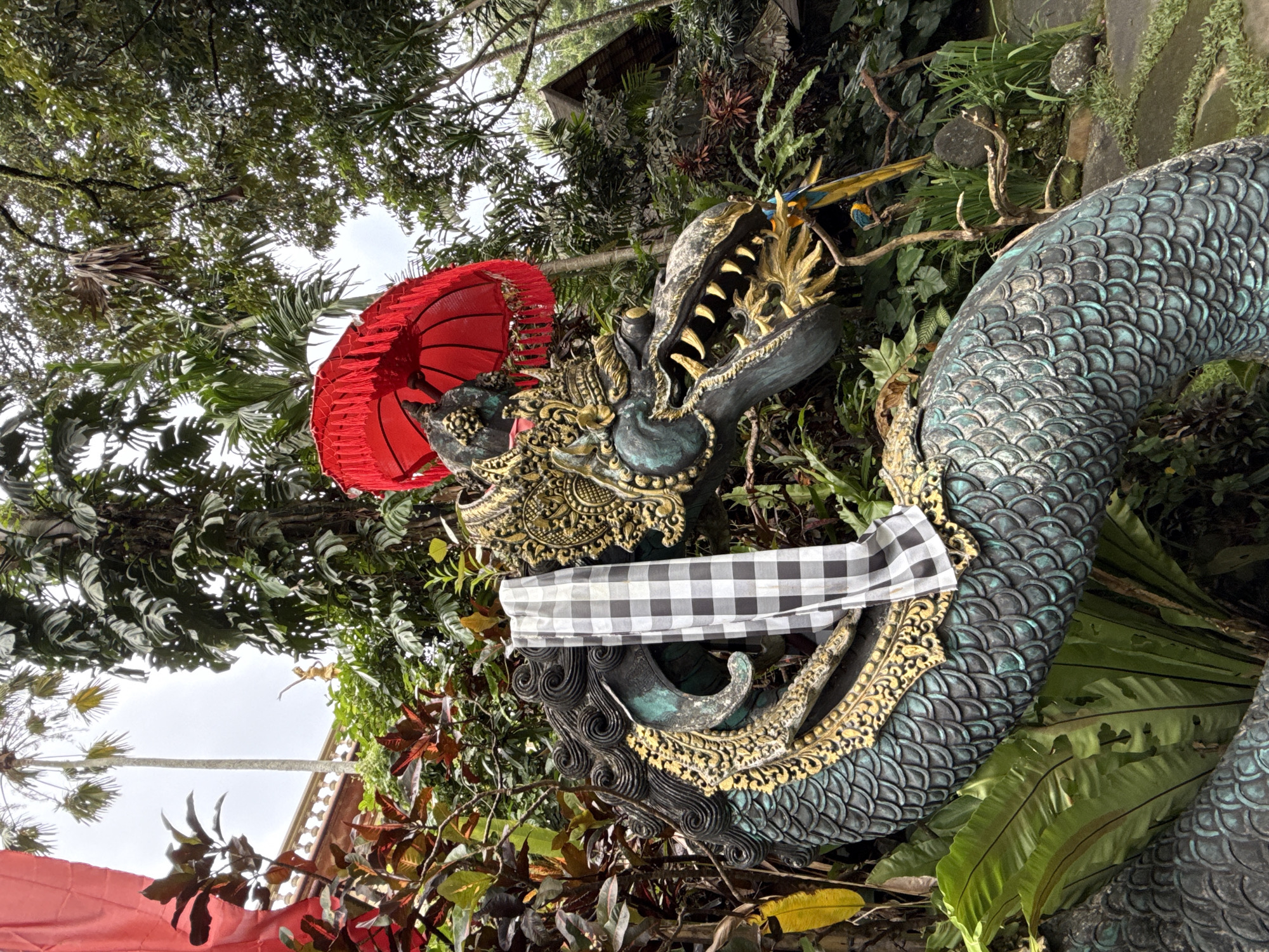 The image shows a detailed statue of a mythical serpent or dragon, likely Naga, common in Balinese culture. The statue is decorated with traditional Balinese elements, including a red umbrella, a checkered black and white cloth (likely a Saput Poleng), and ornate gold accents. The scales of the dragon are rendered in a blue-green hue. The statue is surrounded by lush tropical vegetation, including various types of ferns, bushes, and trees. The location appears to be outdoors, situated within a garden or temple setting. Given the artistic style and cultural elements, it is very likely located in Ubud District, Indonesia. A blue and yellow macaw parrot is also seen perched among the vegetation.