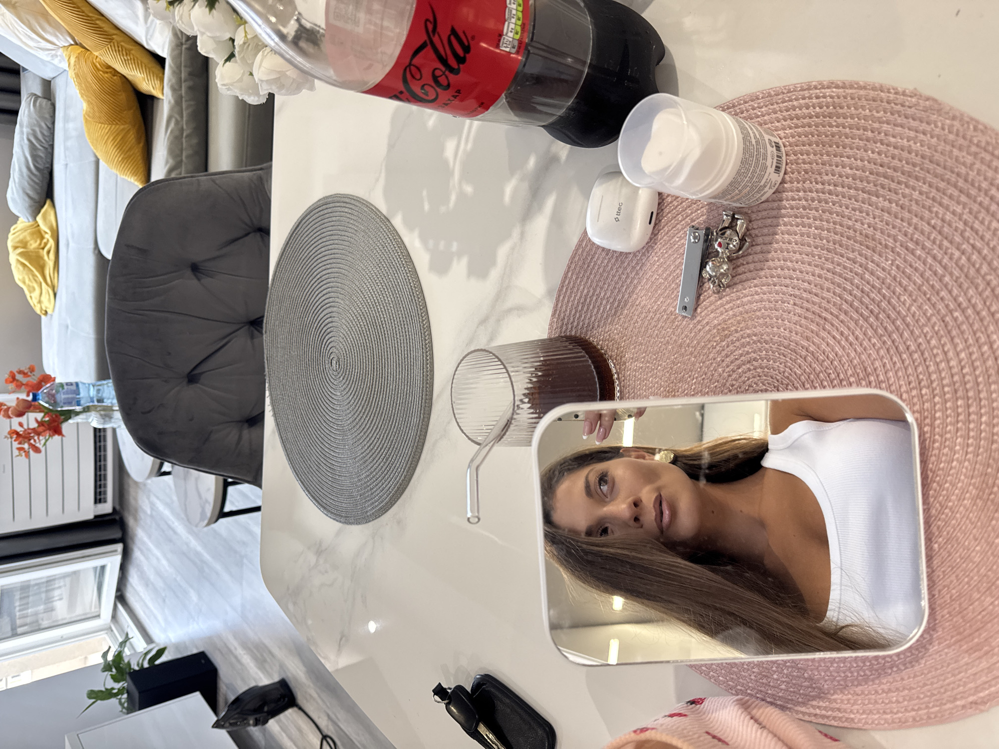 A table in what appears to be an indoor setting holds various items. These include a bottle of Coca-Cola, a white jar, a pair of wireless earbuds, a nail clipper, a drinking glass, a key with a fob, and a hand mirror reflecting the face of a person. The table is adorned with circular placemats, one grey and one pink. A grey velvet chair sits nearby. In the background, there is a radiator, a sofa with yellow pillows, and a plant. The location is not determinable to be in Burgas, Bulgaria from the image.