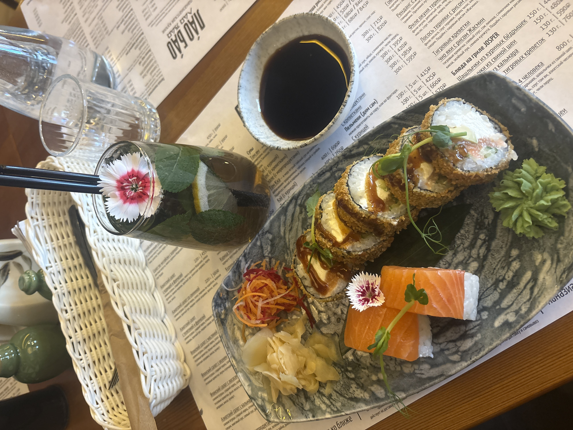 The image captures a food setting, likely within a restaurant in Perm, Russia, given the Cyrillic menu in the background. On a wooden table is a plate of sushi, featuring a variety of rolls and salmon nigiri, garnished with a small pink and white flower and sprouts. There's a small bowl of soy sauce, and a glass containing a beverage, possibly tea or a cocktail, decorated with a slice of lemon, mint leaves, and another pink and white flower. In the background, a glass bottle and additional glassware are visible, along with a white, woven utensil holder. The restaurant's menu is partially visible under the items.