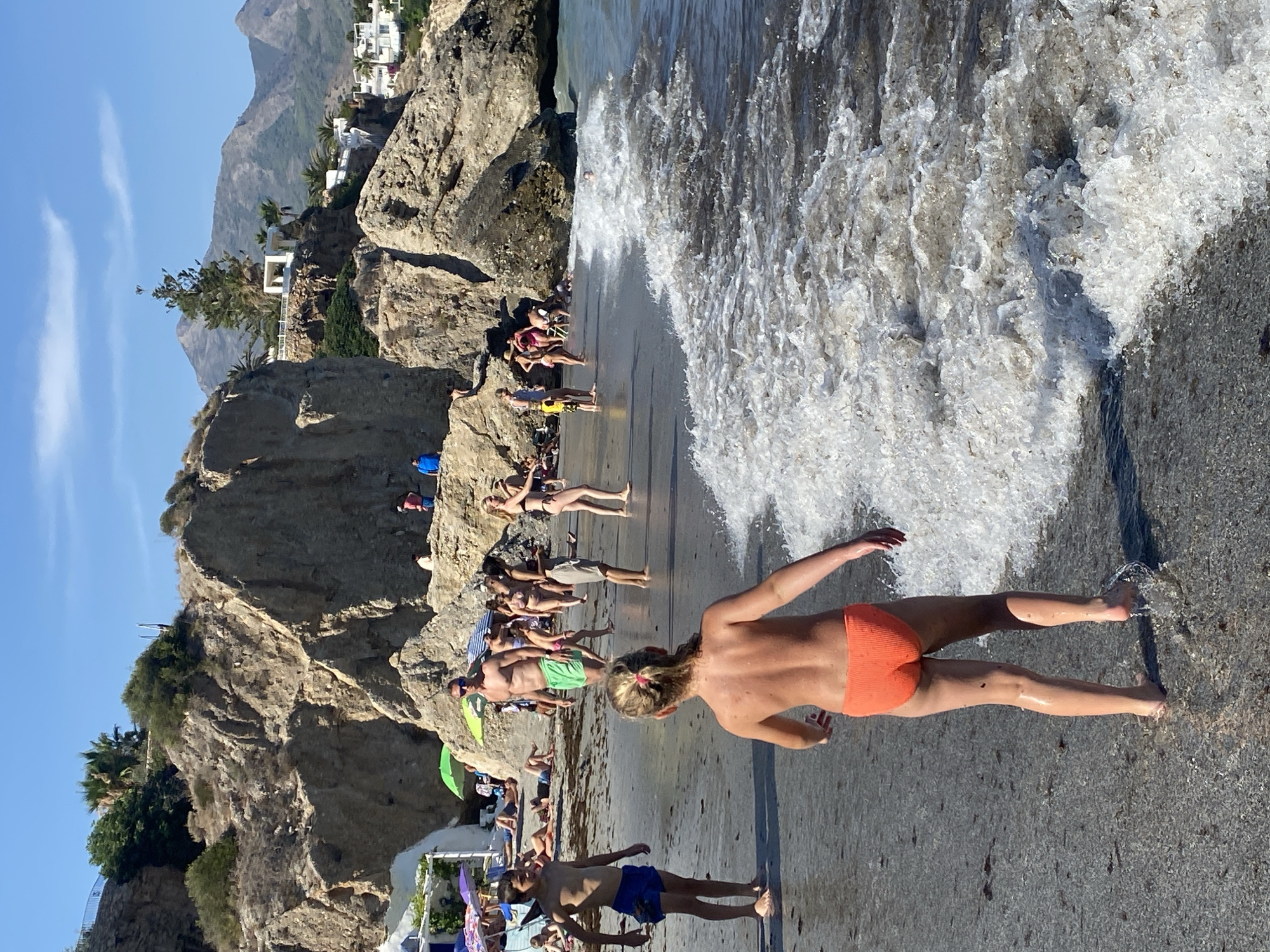 This is a vertical, eye-level shot of a beach in Nerja, Spain. The scene is bathed in bright, natural sunlight, suggesting a warm, clear day. The foreground is dominated by a young person, seen from behind, wearing orange bikini bottoms. Their arms are outstretched as they stand near the water's edge, with waves lapping at their feet and legs.
Further back, a variety of people are spread out on the sandy and rocky shore. Some are sunbathing, some are sitting, and others appear to be standing or walking. Many are dressed in swimwear, highlighting the beach setting. A number of beach umbrellas and towels are visible, indicating a leisure activity.
The background features dramatic, rugged rock formations that rise steeply from the beach. Beyond the rocks, white buildings with red-tiled roofs are nestled on the hillside, hinting at a coastal town. Palm trees and other Mediterranean vegetation dot the landscape, adding to the sense of a vacation destination. The clear blue sky suggests fair weather. The overall atmosphere appears relaxed and summery, with people enjoying a day at the beach. There is no visible text in the image.