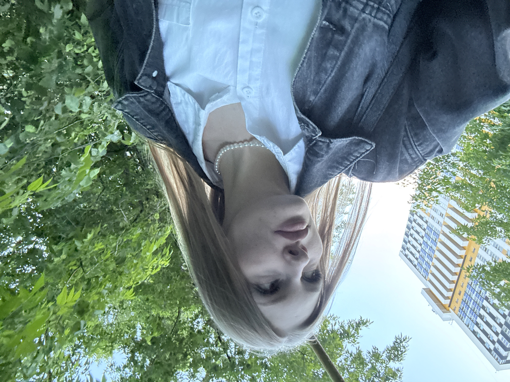 The original image is rotated 90 degrees counter-clockwise. It features a young individual, likely female, in a low-angle shot. Their head is positioned towards the bottom of the frame, and they appear to be looking down towards the camera. The subject has light-colored hair, a neutral expression, and is wearing a white collared shirt, unbuttoned at the top, beneath a dark denim jacket. A pearl necklace is visible around their neck.
The background combines natural and urban elements. Vibrant green foliage from trees or bushes occupies the upper and right portions of the frame. On the bottom-left, against a bright blue sky, a tall, modern residential building with multiple stories, featuring white, yellow, and blue/grey facades, is visible. The bright sky indicates it is daytime with clear weather. The setting suggests an outdoor urban environment, possibly a park or residential area in Perm, Russia. No text is visible in the image.