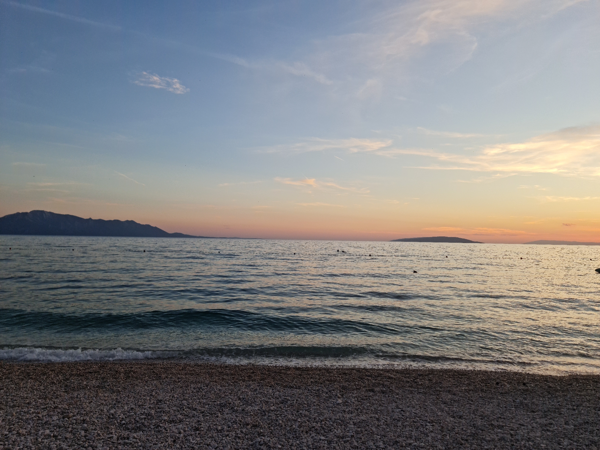 This image depicts a serene beach scene at sunset in Općina Gradac, Croatia. The foreground is dominated by a pebbled shoreline, composed of small, grey, white, and darker stones. Gentle waves with white foam lap at the shore, transitioning into the deeper, dark blue water of the Adriatic Sea. In the middle ground, the sea stretches towards the horizon, with a few dark buoys visible on the surface. The background features a silhouetted mountain range along the left side of the image, with a distinct ridgeline against the fading light. The sky transitions from a pale blue at the top to soft hues of orange and pink near the horizon, indicating the setting sun. Scattered wispy clouds are present in the sky. The overall impression is one of tranquility and natural beauty.