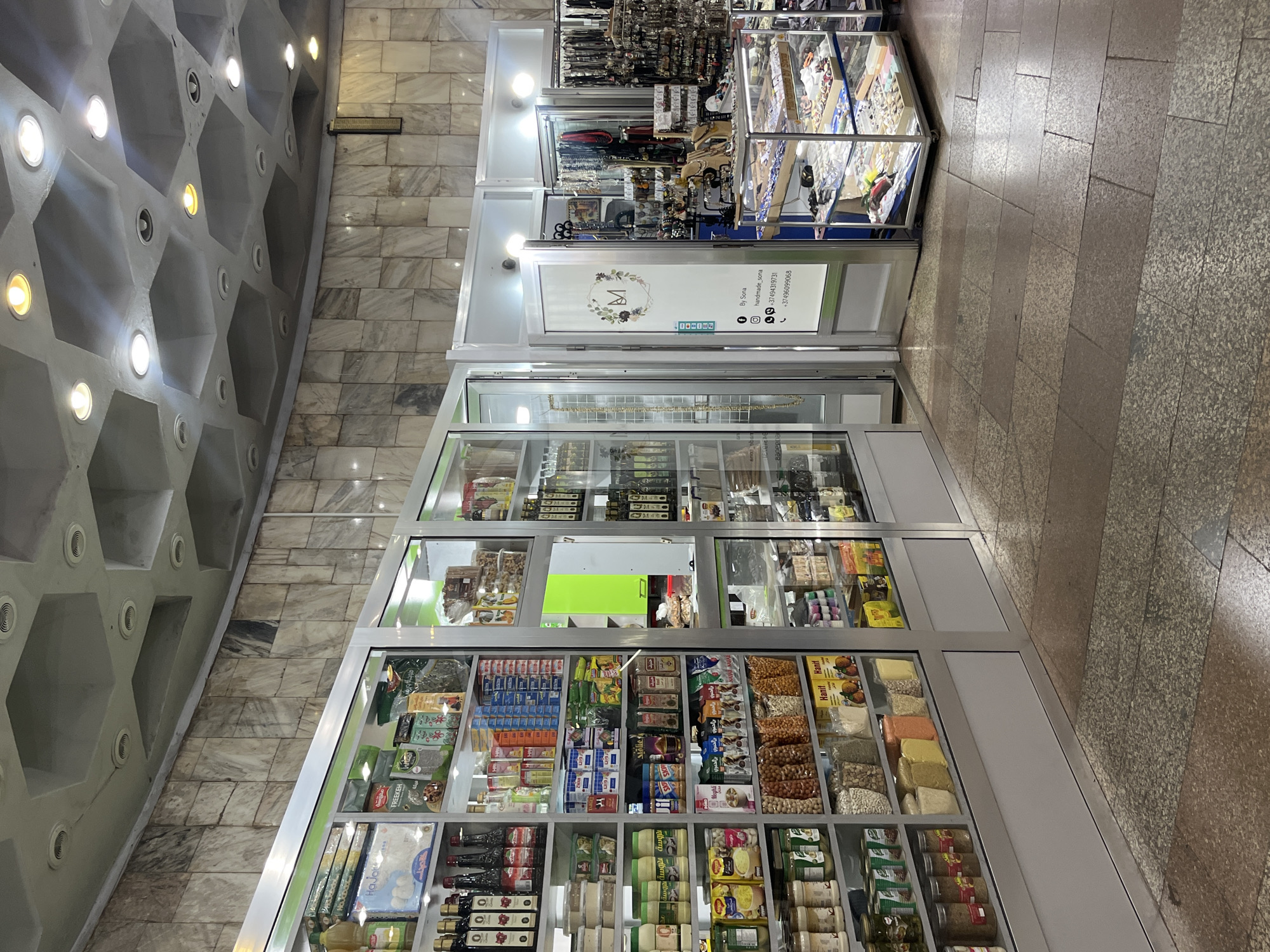 The image shows a shop in Yerevan, Armenia. The shop has multiple display cases filled with various items including jewelry, snacks, and preserved foods. A sign on one of the display cases reads "By Sona handmade_sona +37494319731 +37496099068". The shop appears to be in a mall or shopping center. The floor and ceiling are made of marble.