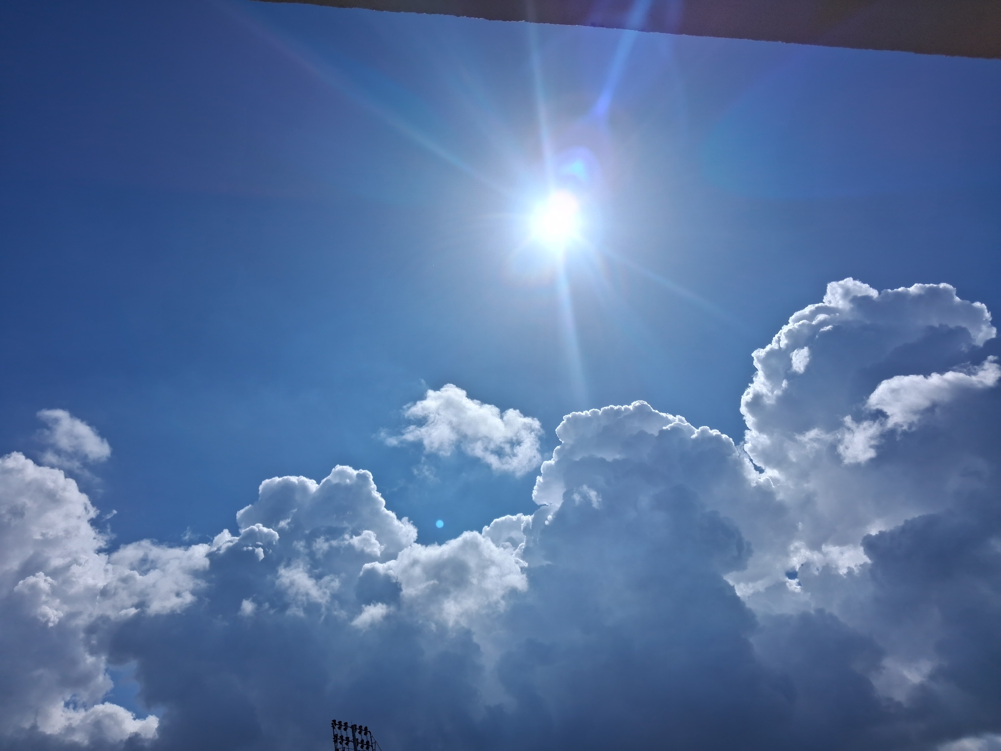 The image presents a bright, sunlit sky filled with cumulus clouds. The sun is high in the sky, emitting a strong glare and lens flare. Below, a scattering of fluffy, white cumulus clouds populates the scene, with darker shadows visible towards the bottom of the frame. The lower edge of the image includes the top of a light tower, which is commonly found in athletic or recreational facilities. The context of Figueira da Foz, Portugal, suggests this scene could be at or near the beach or a local stadium. The image focuses on the natural elements of the sky and clouds.