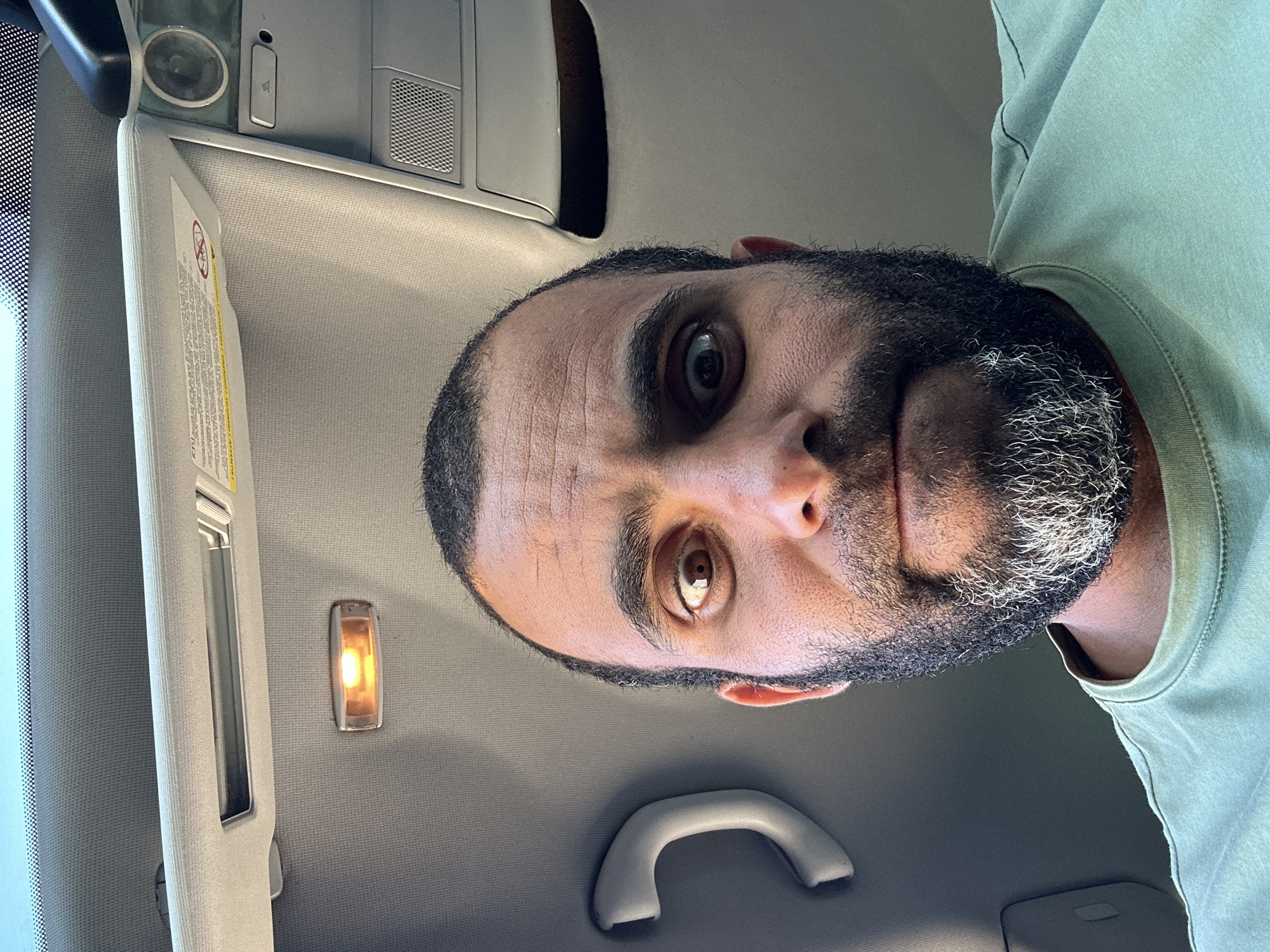 The image is a selfie taken from a low angle inside a vehicle. A man with a dark beard, streaked with gray, is looking directly at the camera with wide, surprised eyes. He is wearing a light green t-shirt. Above him, the interior of the car is visible, including a sun visor with warning labels, an interior light, and a grab handle. The scene appears to be outdoors in daylight, given the lighting conditions. The location of Měčín, Czechia is not discernible from the image itself.