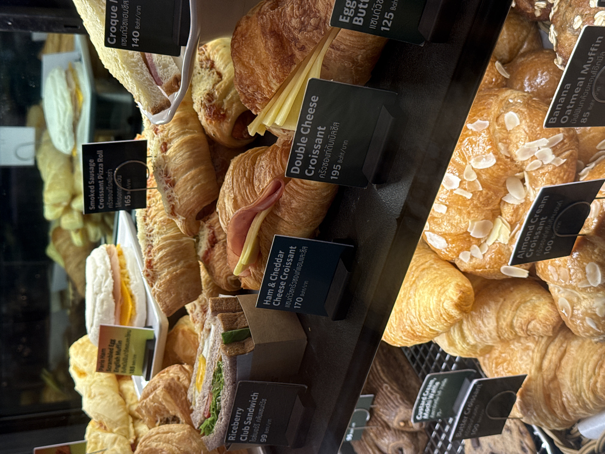 A chilled display case at a Starbucks in Nong Prue Subdistrict, Thailand, showcases a variety of baked goods, including croissants, sandwiches, and muffins. The display case is well-lit, highlighting the textures and colors of the pastries. Among the items featured are a Ham & Cheddar Cheese Croissant, a Double Cheese Croissant, and an Almond Cream Croissant. Also visible are a Riceberry Club Sandwich, a Paris Ham Scrambled Egg English Muffin, and a Smoked Sausage Croissant Pizza Roll. A Croque Monsieur and an Egg Butter croissant are also present. The overall scene suggests a typical retail environment for takeaway food items.