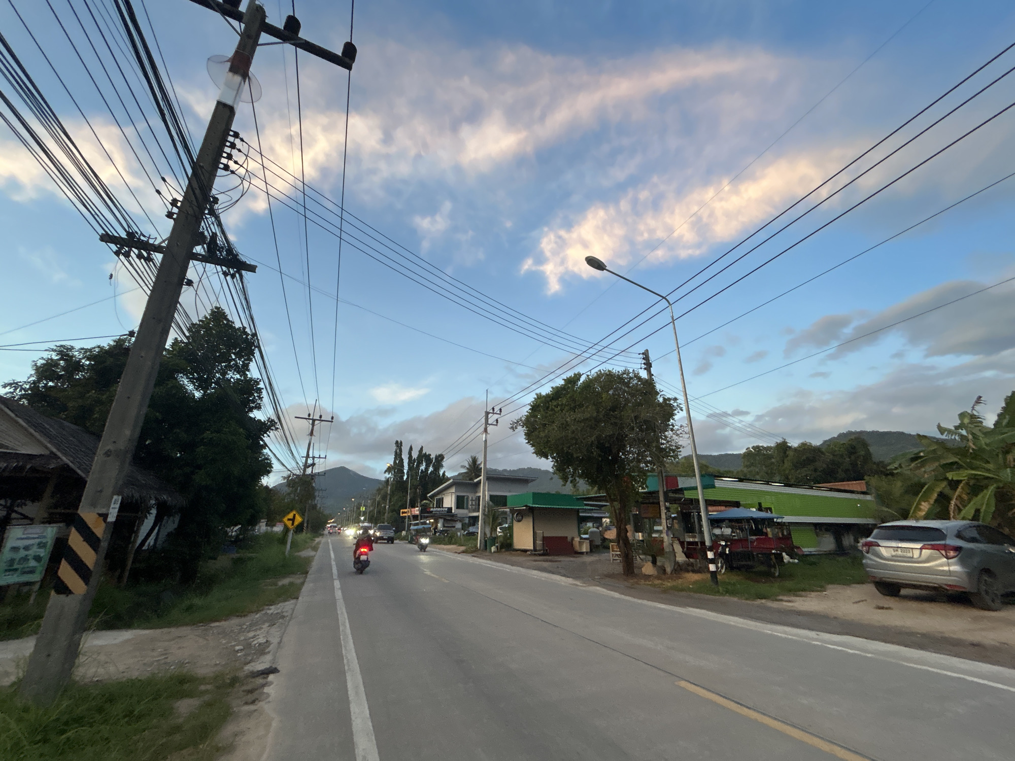 The image shows a street scene in ตำบลเกาะพะงัน, Thailand. The road is gray asphalt with a yellow line marking the edge. Two motorcycles are driving on the left side of the road in the distance. A light pole is leaning to the left side of the frame, with numerous wires extending from it. Buildings are present on the right side of the road, including a green structure with a blue awning and a gray car parked nearby. The sky is blue with scattered clouds. Green vegetation is on both sides of the road, including trees and shrubs. A mountain is visible in the background.
