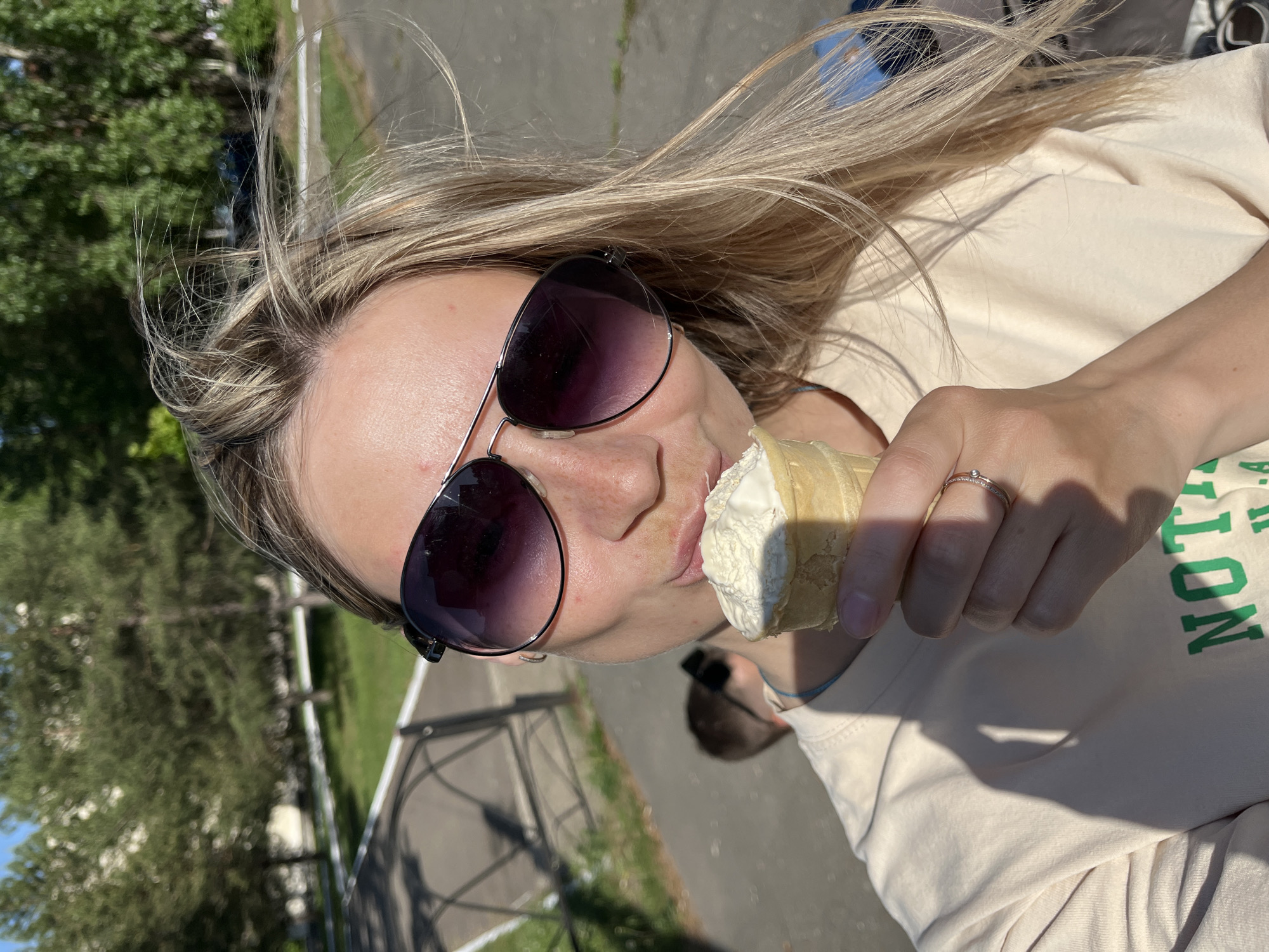 A woman is eating an ice cream cone. She has blonde hair and is wearing sunglasses. She is wearing a light-colored t-shirt with green lettering. She is also wearing a ring on her finger. The background appears to be a park or outdoor area. It is hard to identify the specific location in Chaikovsky, Russia, without additional information.