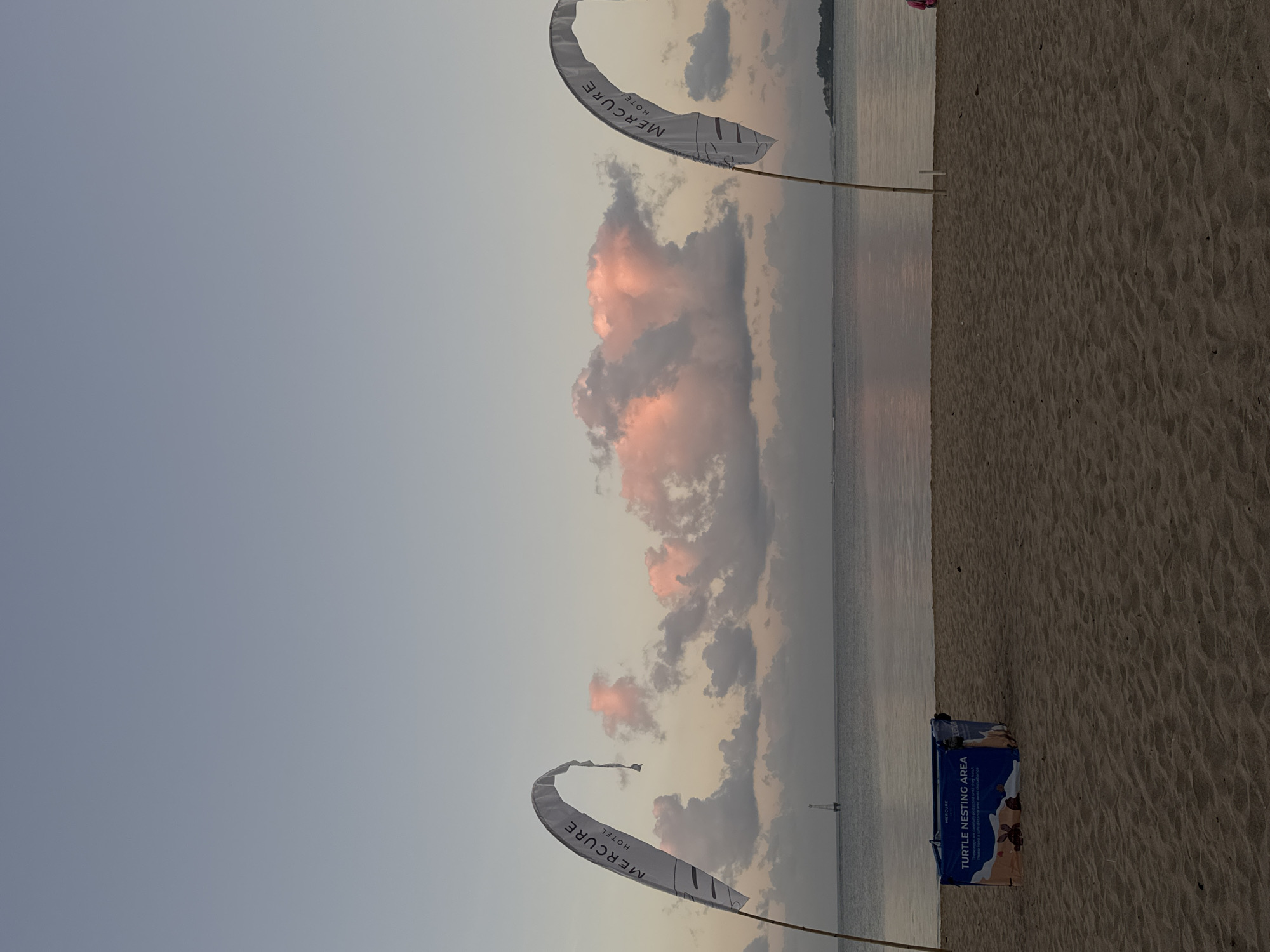 The image shows a beach scene at twilight, likely in Denpasar, Indonesia. The sky is a gradient of soft blue and pink hues, with several fluffy clouds tinged with pink from the setting sun. The ocean is calm and reflects the colors of the sky. In the foreground, there is a sandy beach. On the left side of the beach, there is a white flag with the "MERCURE HOTEL" logo printed on it, attached to a bamboo pole. To the right of this flag, there is a blue and orange box labeled "TURTLE NESTING AREA" with graphics of turtles. Further down the beach, on the right side, another white "MERCURE HOTEL" flag is visible. In the distance, a small lighthouse or buoy can be seen in the water, and a faint outline of land is visible on the horizon. The overall atmosphere is serene and peaceful.