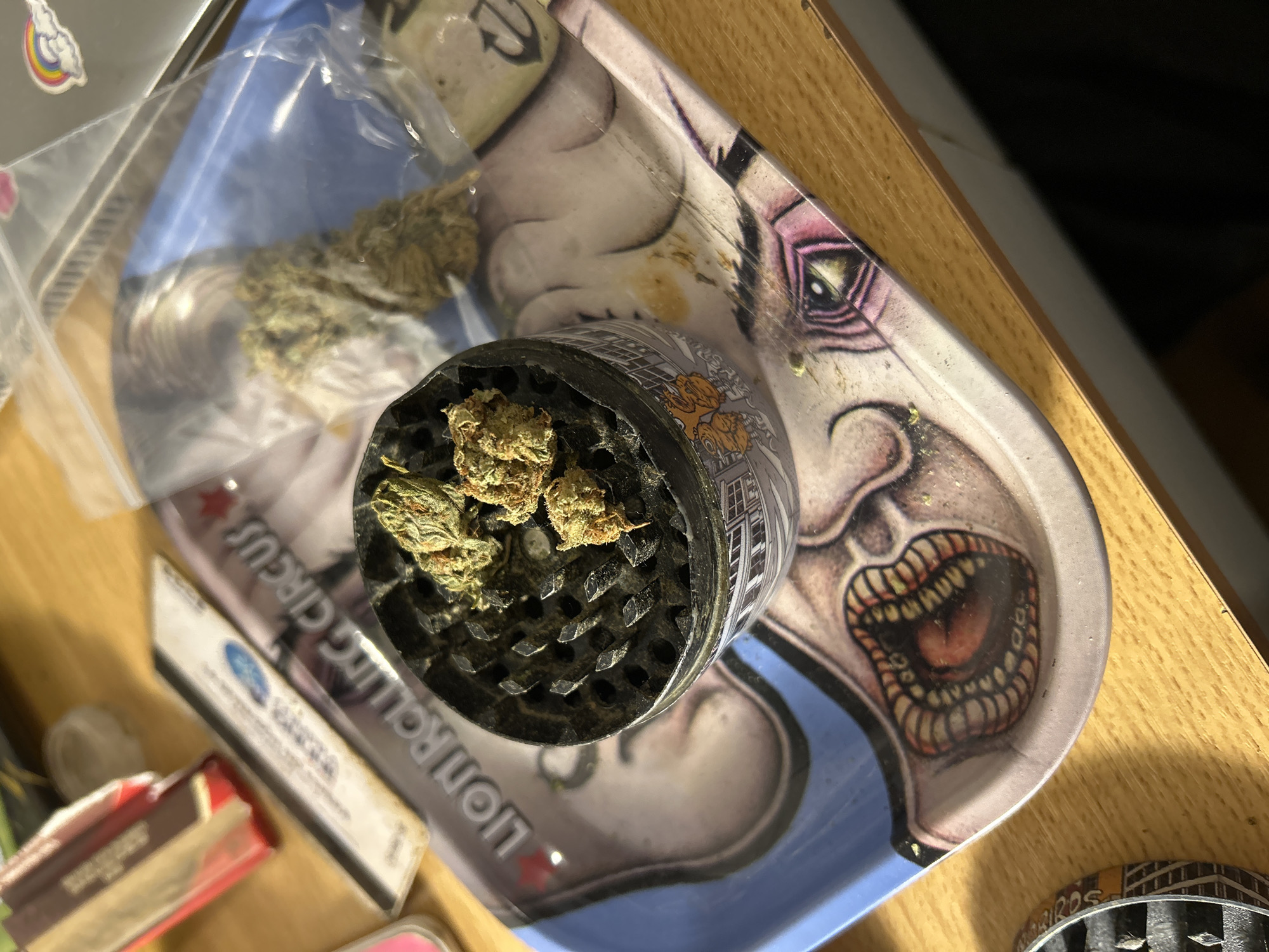 On a wooden surface, a rolling tray with a graphic of a monster's face is visible, bearing the inscription "LIONROLLING CIRCUS." A metal grinder containing marijuana buds sits atop the tray. Beside the tray are rolling papers and other associated items. The location is not discernible from the image.
