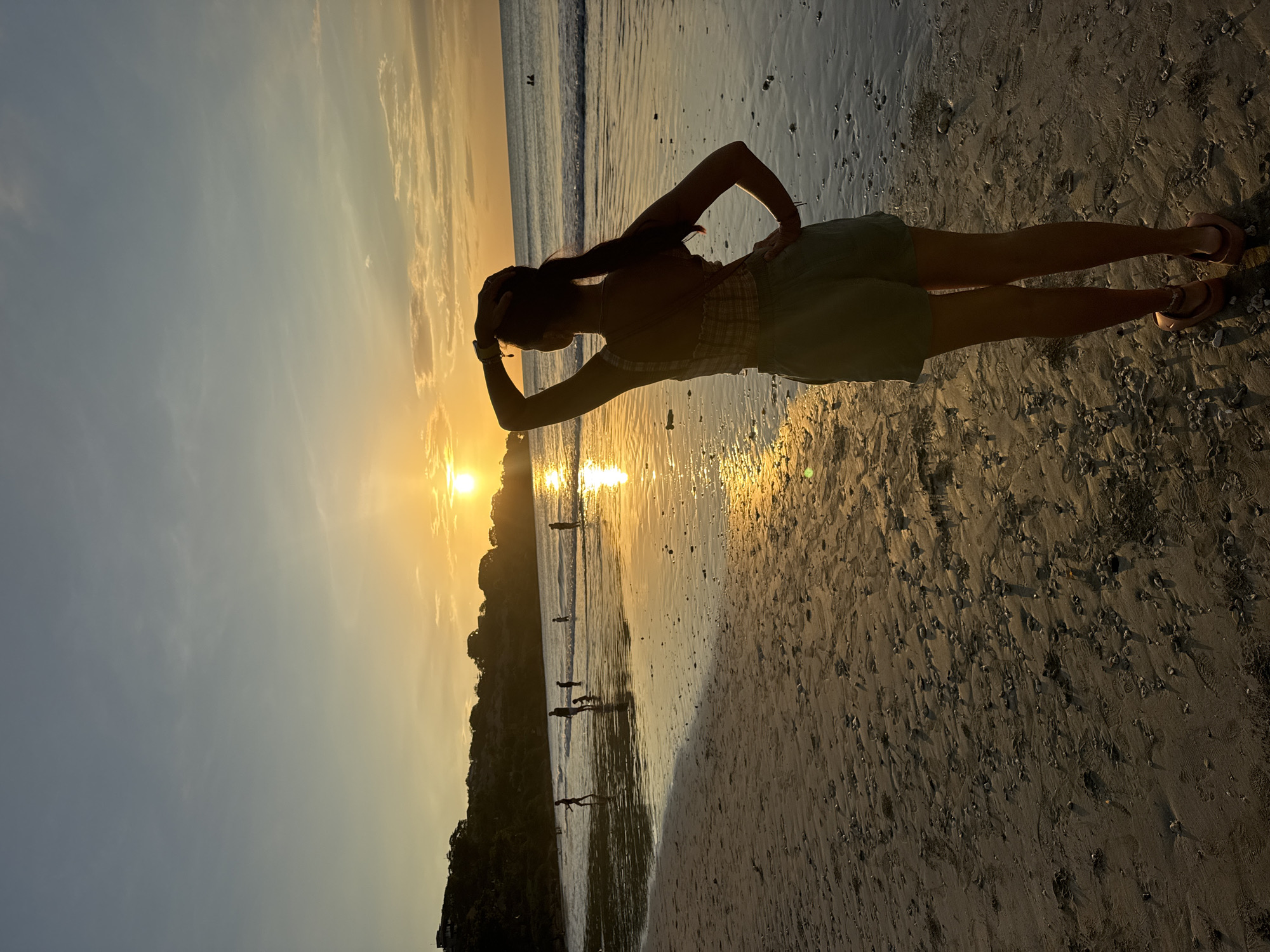 A silhouetted female figure stands on a sandy beach at sunset in Kuta Selatan, Indonesia. The woman, wearing a light-colored sundress and sandals, is positioned facing the ocean with her left hand on her head and her right hand on her hip. The sun is setting over the ocean, casting a golden glow on the sand and water. Several other silhouetted figures are visible walking along the beach near the water's edge. A land formation covered in trees can be seen to the left, near the sunset. The sand has a grainy texture and is wet near the ocean.