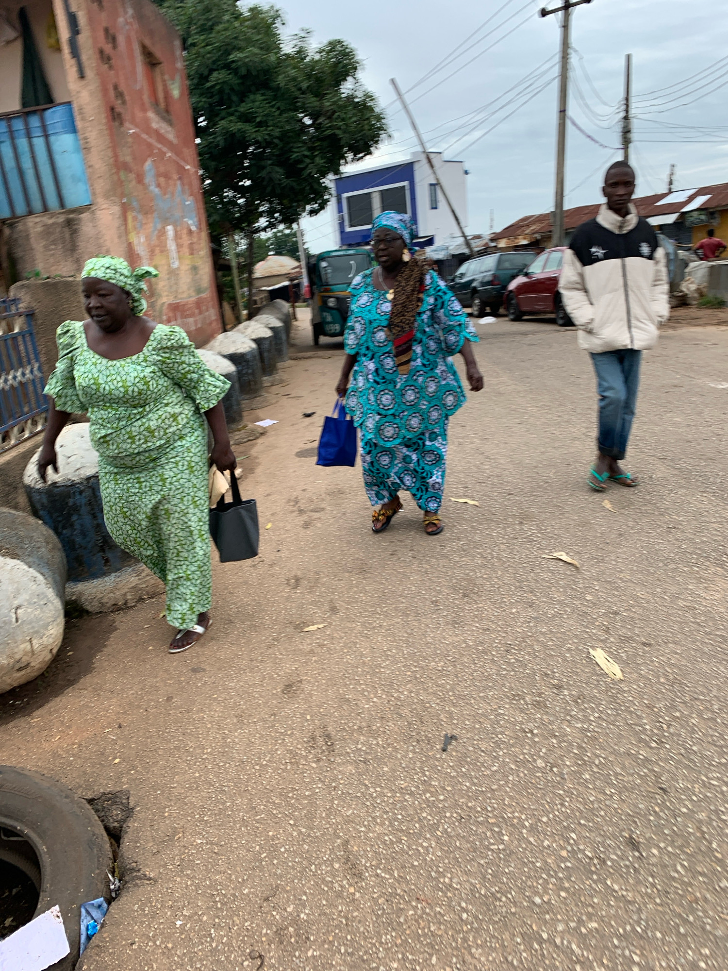 In Jos, Nigeria, a street scene unfolds with three individuals walking on a paved road. On the left, a woman in a green patterned dress and headscarf carries a dark tote bag in her right hand. She wears white sandals. To her right, a woman in a blue and teal patterned dress and headscarf walks with her head slightly turned. She wears sandals and a patterned scarf around her neck. Further to the right, a young man in a black and white puffer jacket and blue jeans walks facing forward, wearing dark sandals. The environment includes a red-brick building with a blue-barred window on the left, a row of cylindrical concrete barriers, a utility pole with wires overhead, and parked vehicles in the background. A three-wheeled vehicle, possibly an auto-rickshaw, is visible in the middle ground. The ground is asphalt, with scattered dry leaves and debris. The sky is overcast.