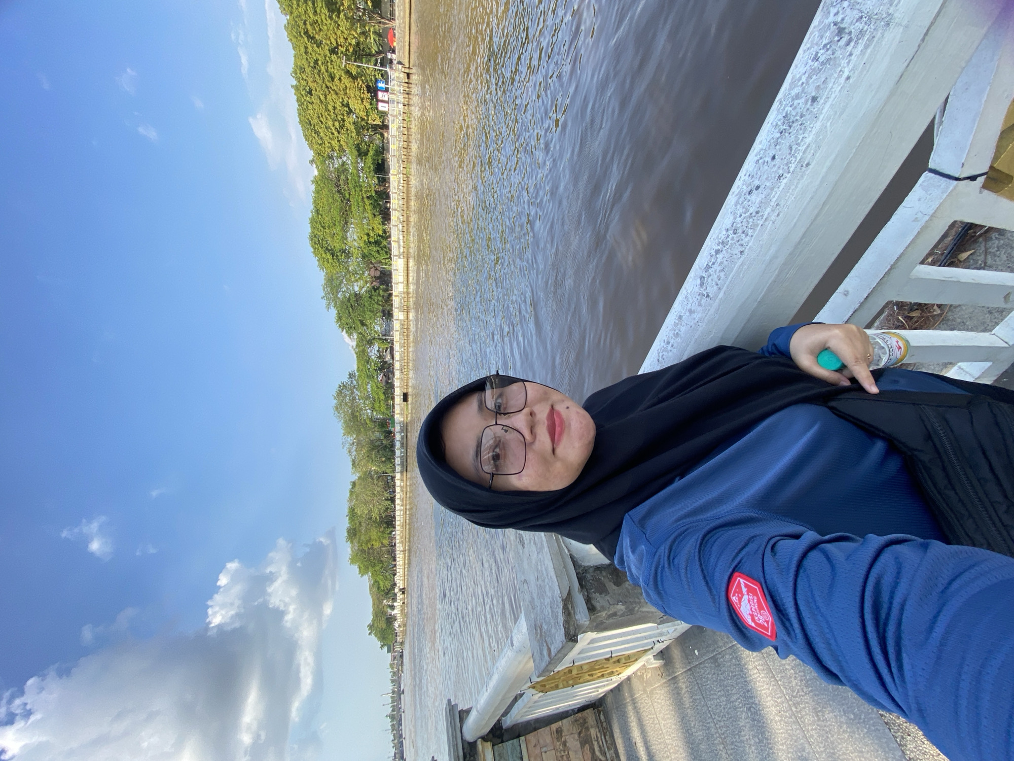 A woman wearing a black hijab and a blue long-sleeved shirt is taking a selfie on a bridge overlooking a river. She is wearing glasses and a red patch with white text and a red mountain logo is visible on her left sleeve. In her right hand, she is holding a clear water bottle with a green cap. The river is wide and brown, with gentle ripples on its surface. Along the riverbank, there is a white railing with gold accents, and beyond that, a row of trees and buildings can be seen under a blue sky with scattered white clouds. The scene appears to be in a city, with the presence of a bridge and buildings suggesting an urban environment. The lighting indicates it is daytime, likely late afternoon given the angle of the sun casting shadows. The overall atmosphere is relaxed and casual, consistent with a personal photograph. This location is likely the Martapura River in Banjarmasin, Indonesia, a city known for its rivers and bridges.