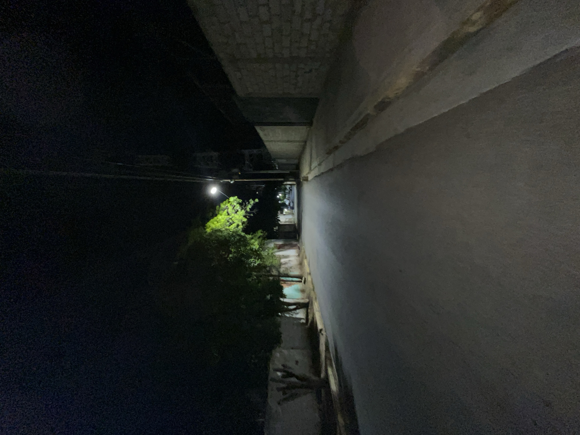 The image depicts a street scene at night in Santa Ana, Venezuela. A paved road runs through the center of the frame, bordered on the left by a low, light-colored wall and trees, some of which are illuminated by a streetlamp. On the right, a sidewalk runs alongside a similar low wall constructed of concrete blocks. The street is empty, with no people or vehicles visible. The sky is dark and appears to be clear.