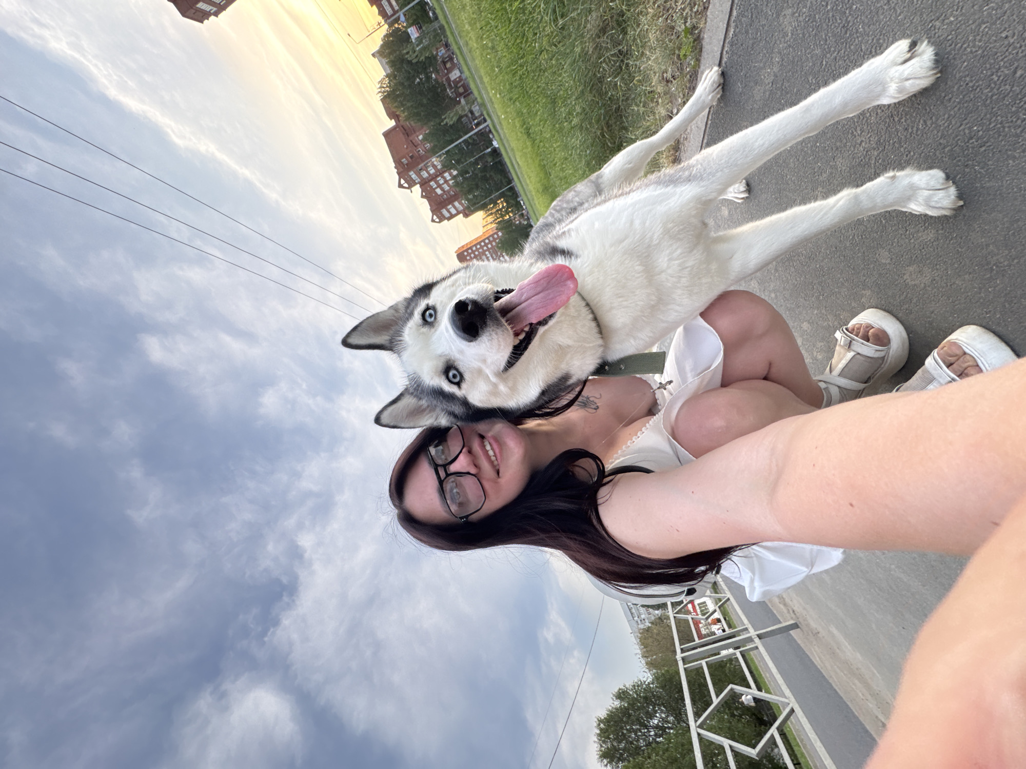 A medium shot captures a woman and a dog on a sidewalk in an urban setting. The woman, with dark hair, glasses, and a white dress, holds the camera at arm's length, seemingly taking a selfie. The dog, a husky with white and grey fur, is positioned beside her head, tongue lolling out. The sidewalk is grey, and patches of green grass and trees are visible along the side. In the background, there are buildings with red brick facades. The sky is a mix of clouds and sunlight. A fence is seen. The location is the city of Unknown, Unknown.