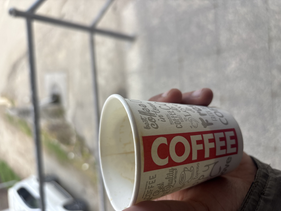 The scene captures a close-up, downward-angled perspective of a human hand holding a white disposable paper coffee cup. The cup features a prominent vertical red bar with "COFFEE" in white capital letters on one side, and various coffee-related words and icons in grey text on the remainder of its surface, including phrases like "best coffee in town" and "morning coffee." The cup's interior shows residue consistent with a consumed beverage, indicating it is partially empty. The hand holding the cup displays visible skin texture and creases. The background is significantly blurred, depicting an outdoor environment. Immediately behind the cup, a dark, thin metal railing runs horizontally across the frame. Below and beyond the railing, a light-colored concrete or paved surface is visible, alongside which a portion of a white passenger vehicle is discernible on the left. Faint green foliage or grass appears next to the paved area in the distance. The overall impression is an individual holding a coffee cup outdoors from an elevated position. This scene is located in the city of Lehvaz, Armenia.