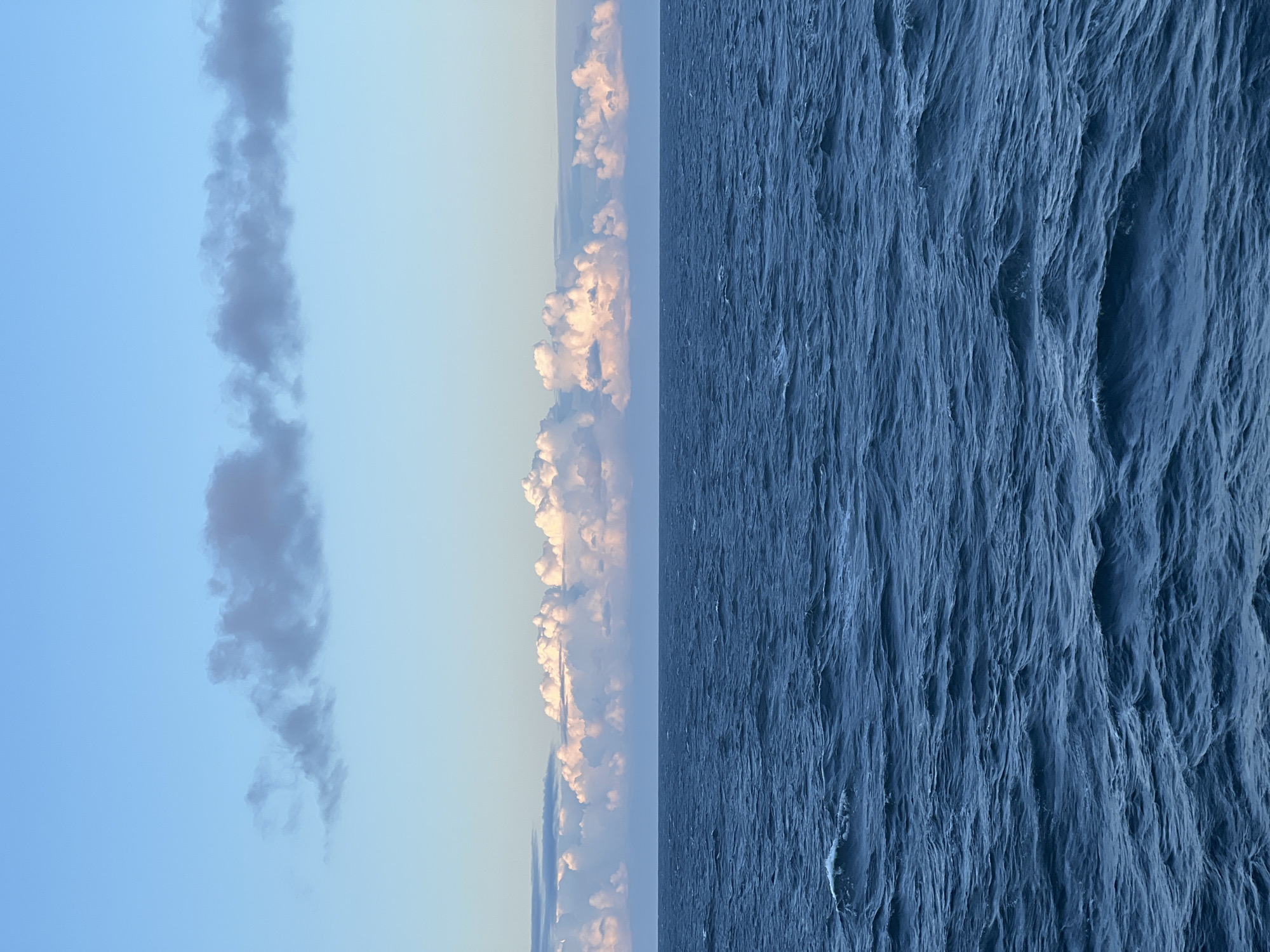 The image depicts a vast expanse of dark blue, choppy ocean under a pale blue sky. Several layers of clouds are visible. A thin band of hazy, gray clouds stretches horizontally across the upper portion of the sky, while below it, a more prominent line of puffy, white clouds, tinged with pink and orange, sits just above the horizon. The rough texture of the waves suggests a breezy or windy condition. The scene is devoid of any human presence or man-made structures, presenting a purely natural landscape. The location is identified as Unknown, Poland.